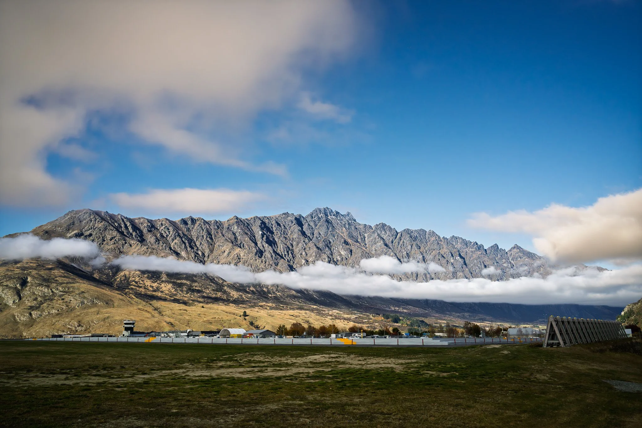 Mountain range with rocky peaks, low clouds, and a flat ground area in the foreground, under a partly cloudy blue sky.