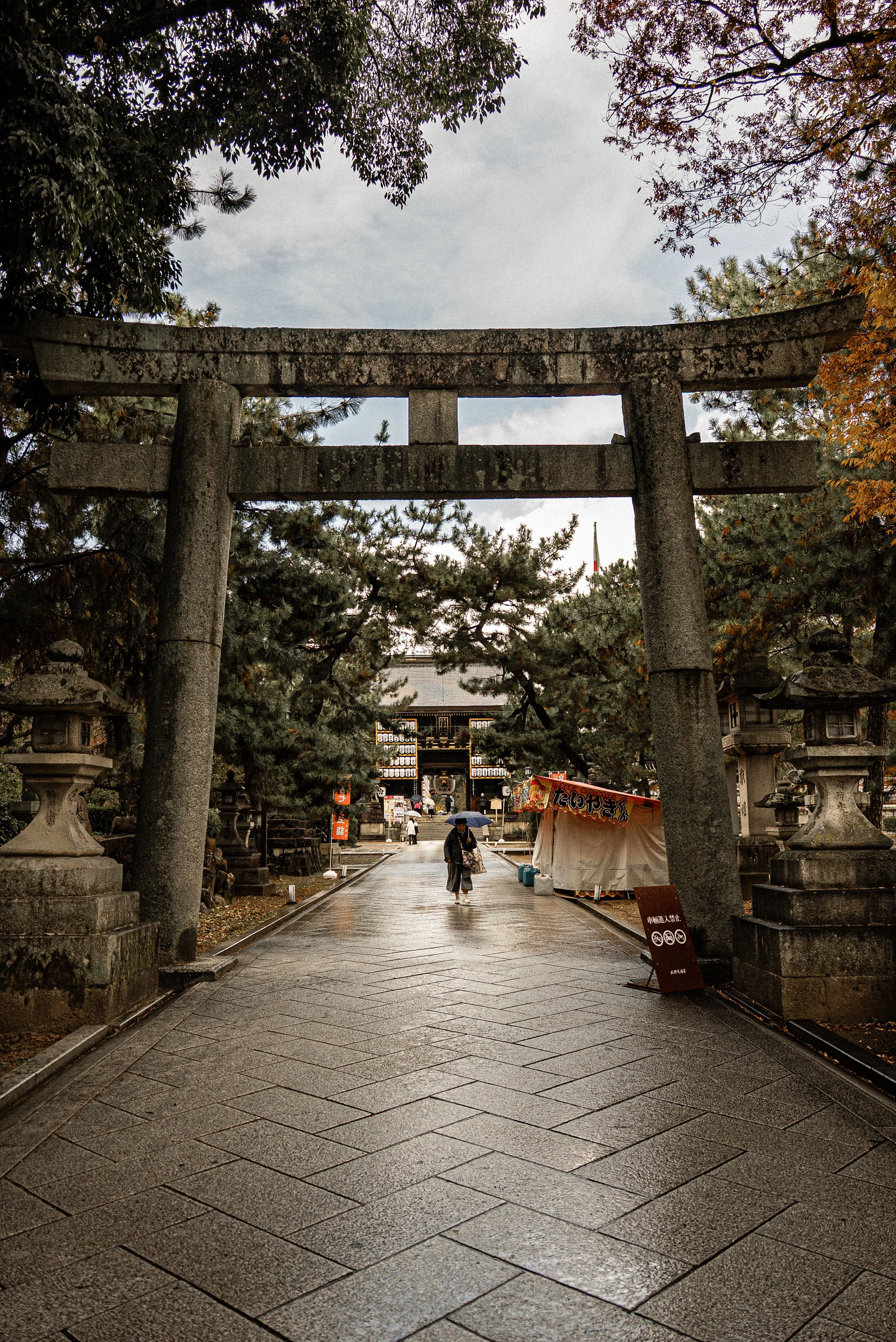 A large traditional Japanese torii gate made of stone, standing at the entrance of a shrine, surrounded by trees and a gray sky. A person holding an umbrella walks along the wet stone-paved path underneath.
