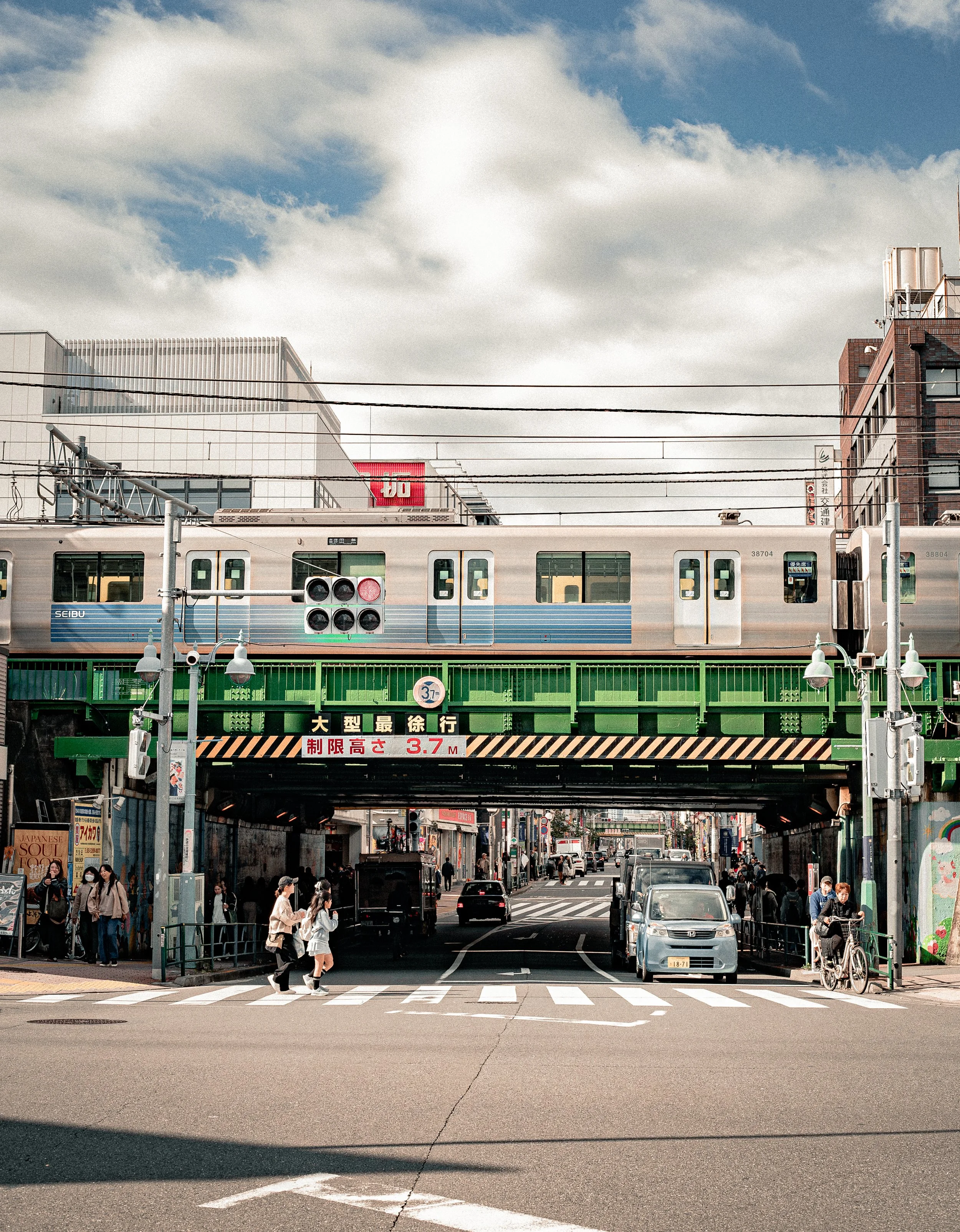 A busy city street scene with a train passing overhead on an elevated track, with pedestrians walking on the crosswalk below, cars on the road, and buildings lining the street under a partly cloudy sky.