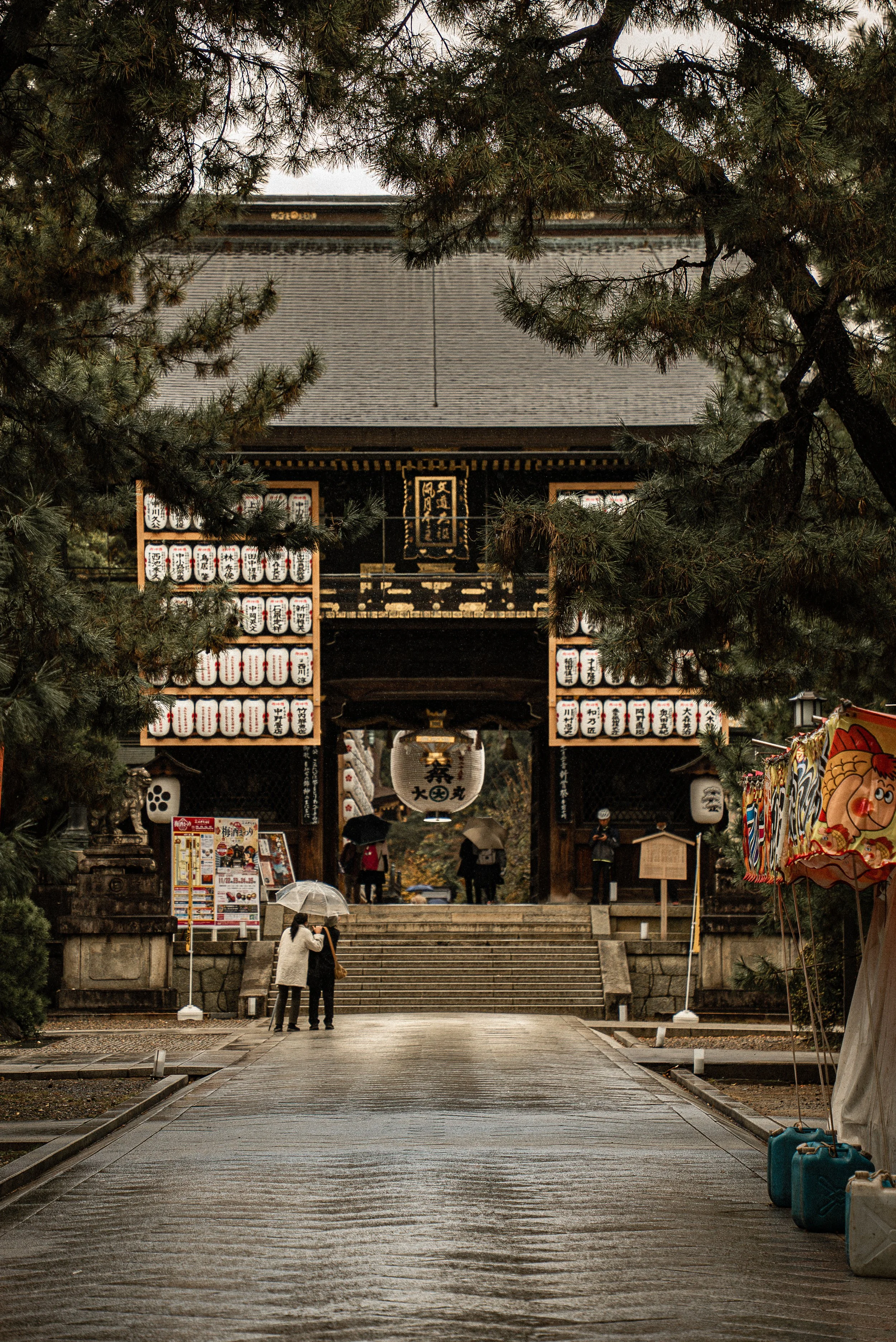 People walking towards the entrance of a traditional Japanese temple, surrounded by trees and lanterns, on a rainy day.