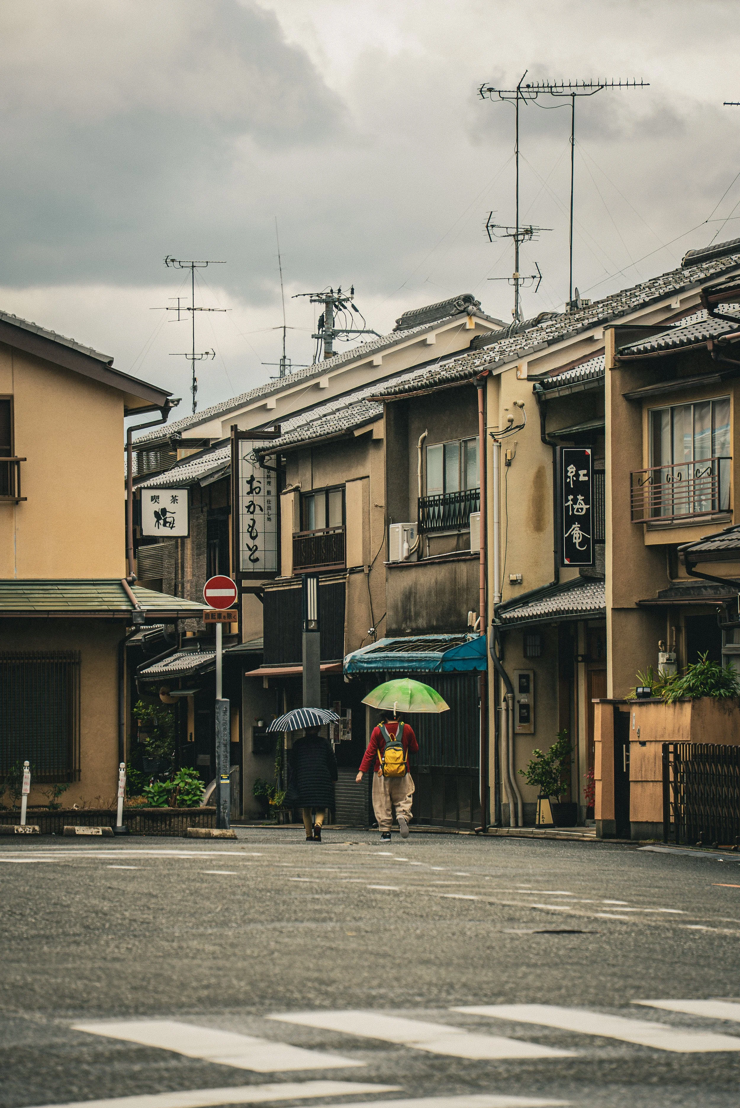 A street scene in Japan with two pedestrians walking with umbrellas, surrounded by traditional buildings with signs, and an overcast sky with numerous TV antennas on rooftops.