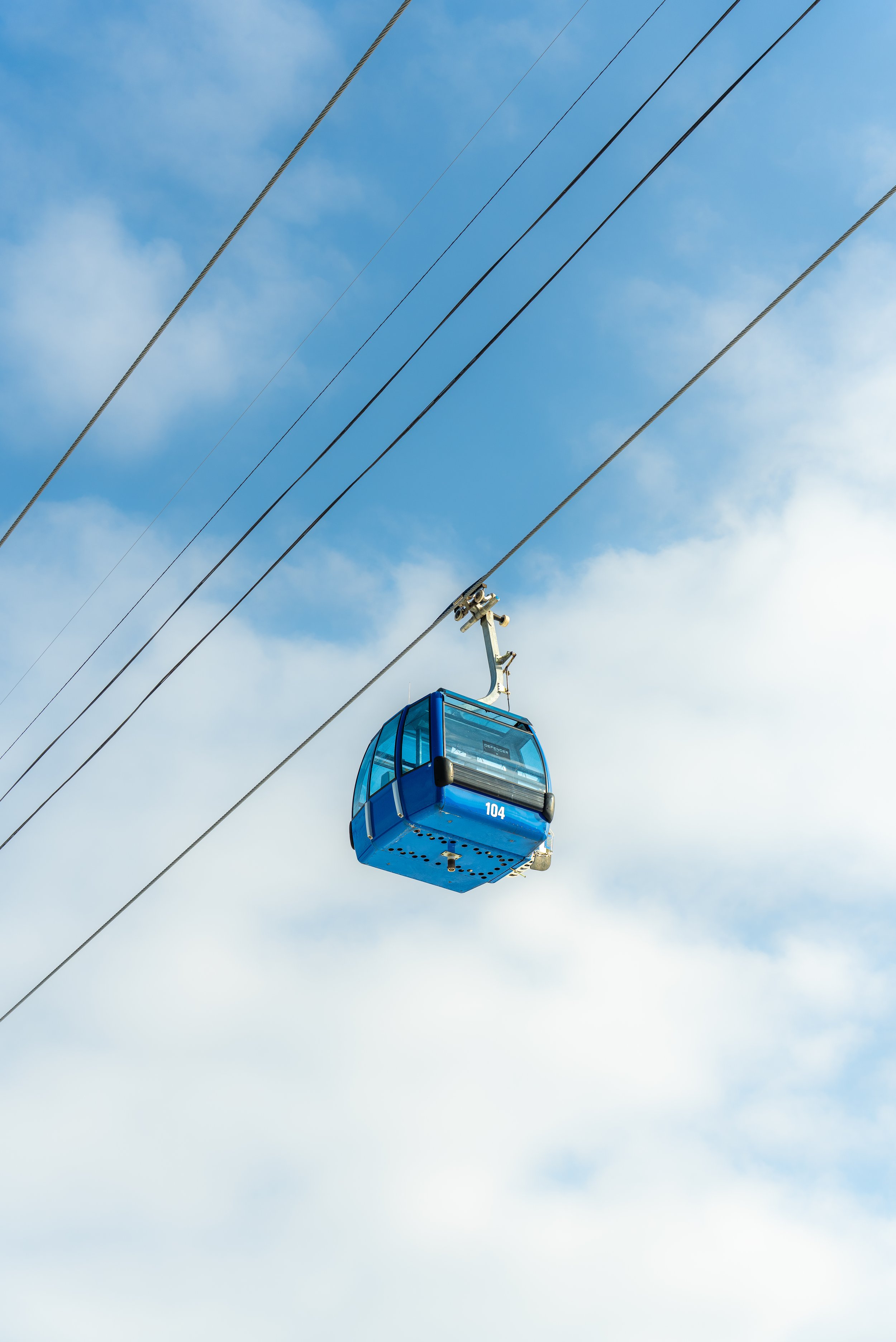 A blue cable car labeled 104 moving through the sky with clouds and electrical wires overhead.