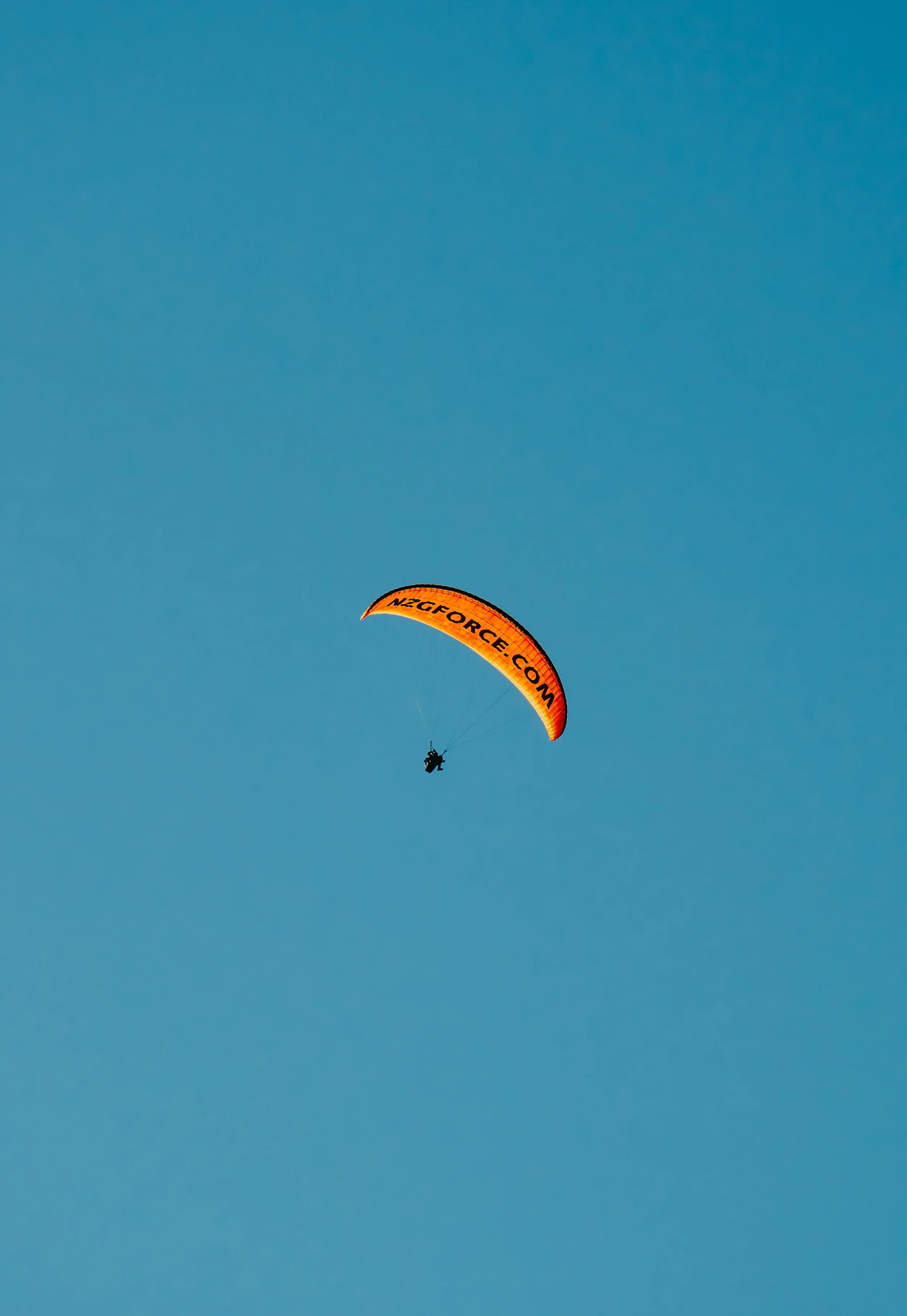A person paragliding with an orange and yellow parachute over a clear blue sky.