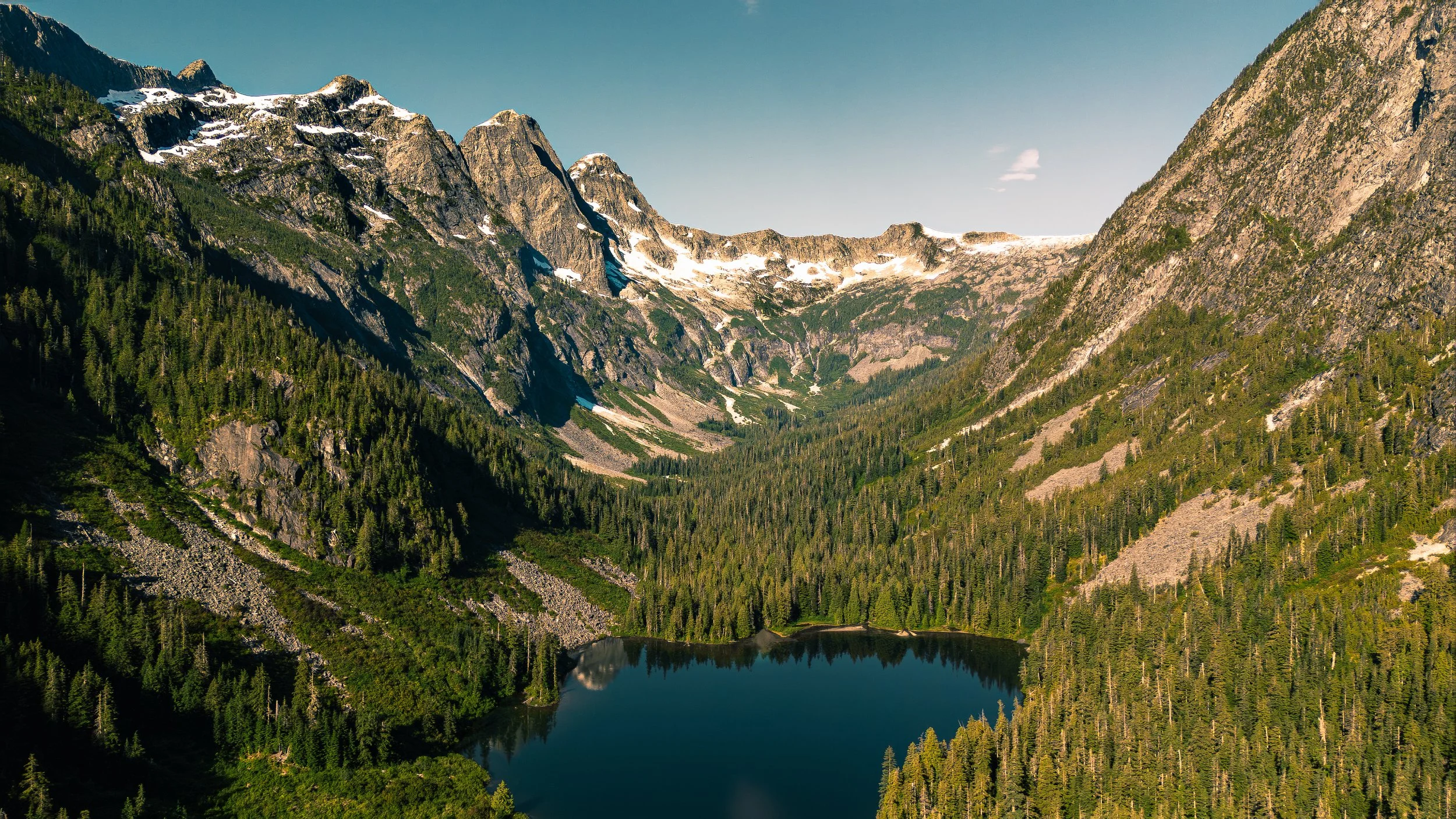 A scenic view of a mountainous landscape with rocky peaks, snow patches, dense green forests, and a calm lake reflecting the mountains under a clear blue sky.