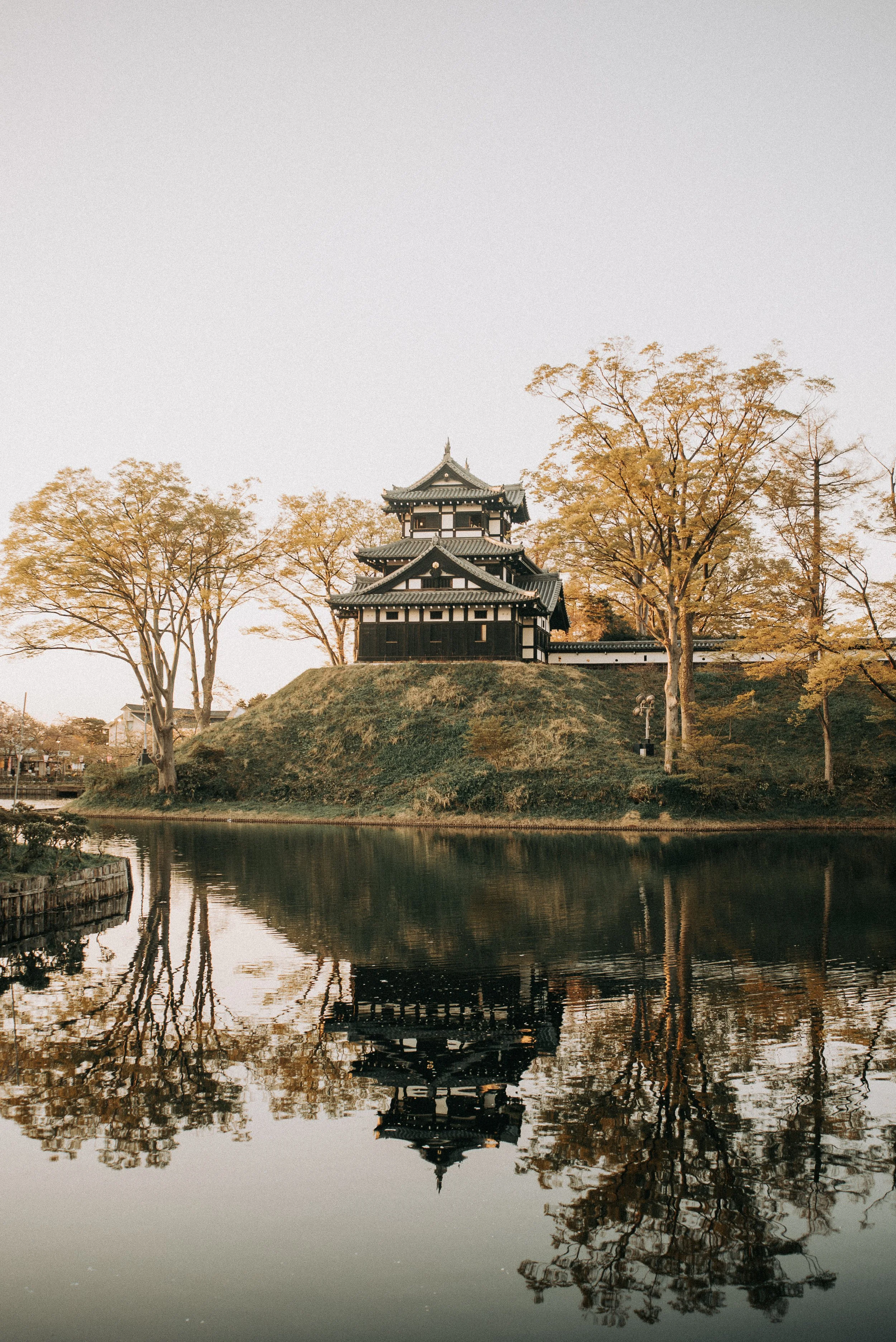 A traditional Japanese castle on a hill, surrounded by trees with autumn foliage, reflected in a calm body of water.