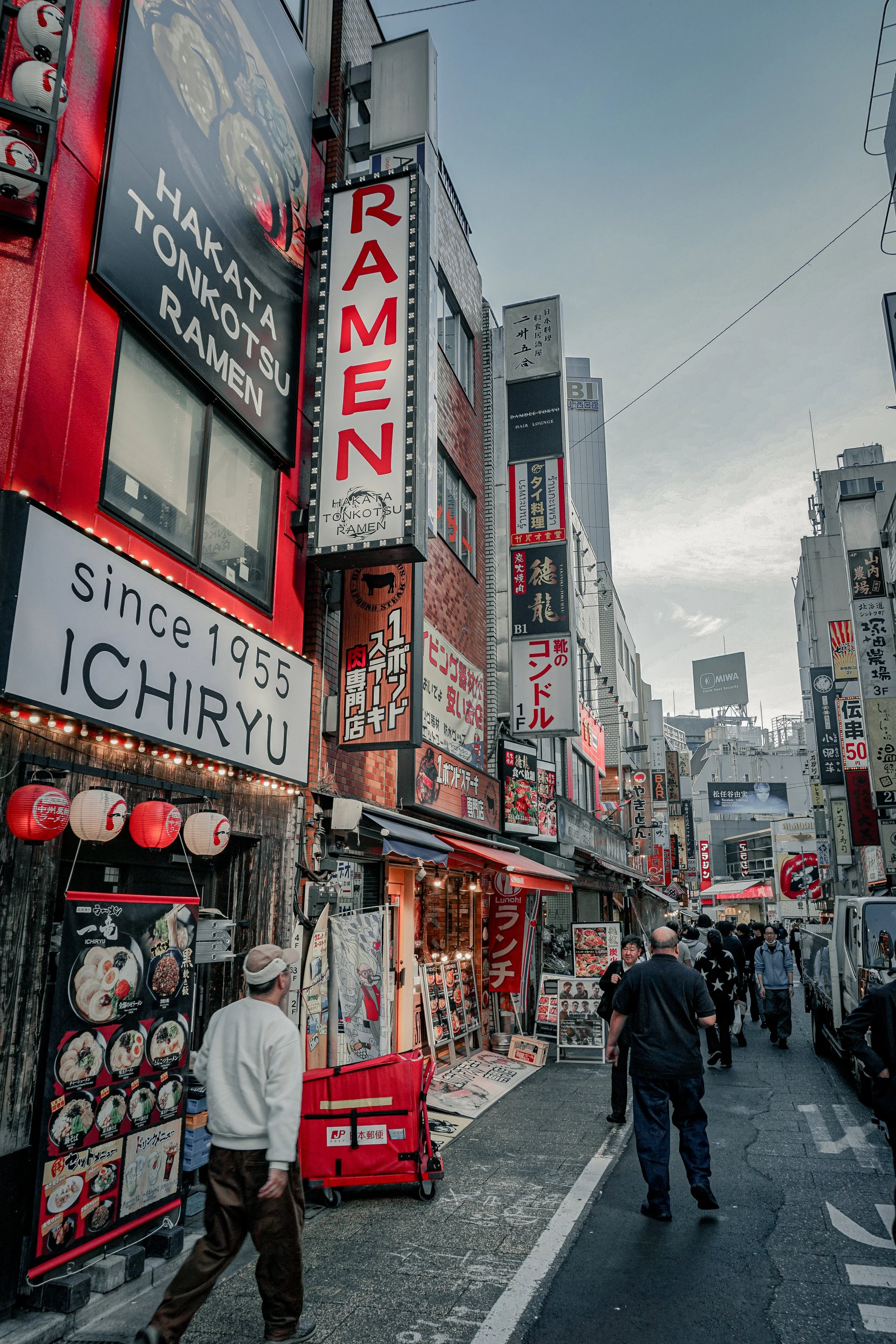 City street in Japan with signs in Japanese and English. People walking, restaurants and shops visible. Red and black color theme, lanterns hanging outside the restaurant.
