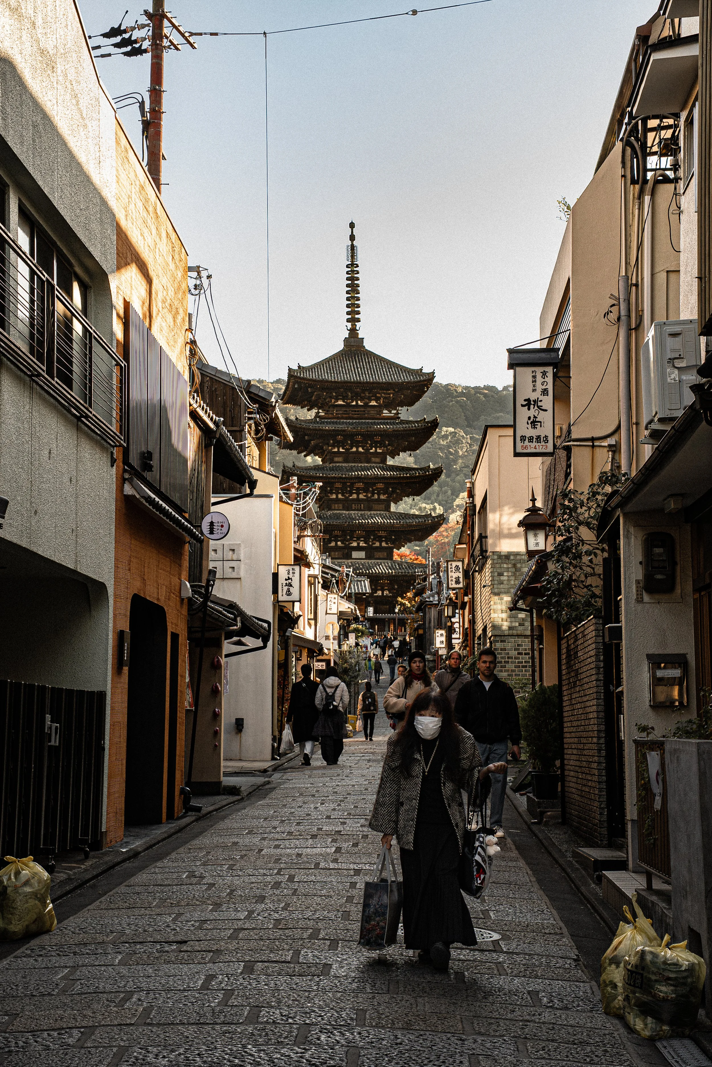 Pedestrians walking on a narrow street in Kyoto, Japan, with traditional buildings on both sides and a pagoda-style temple in the background.