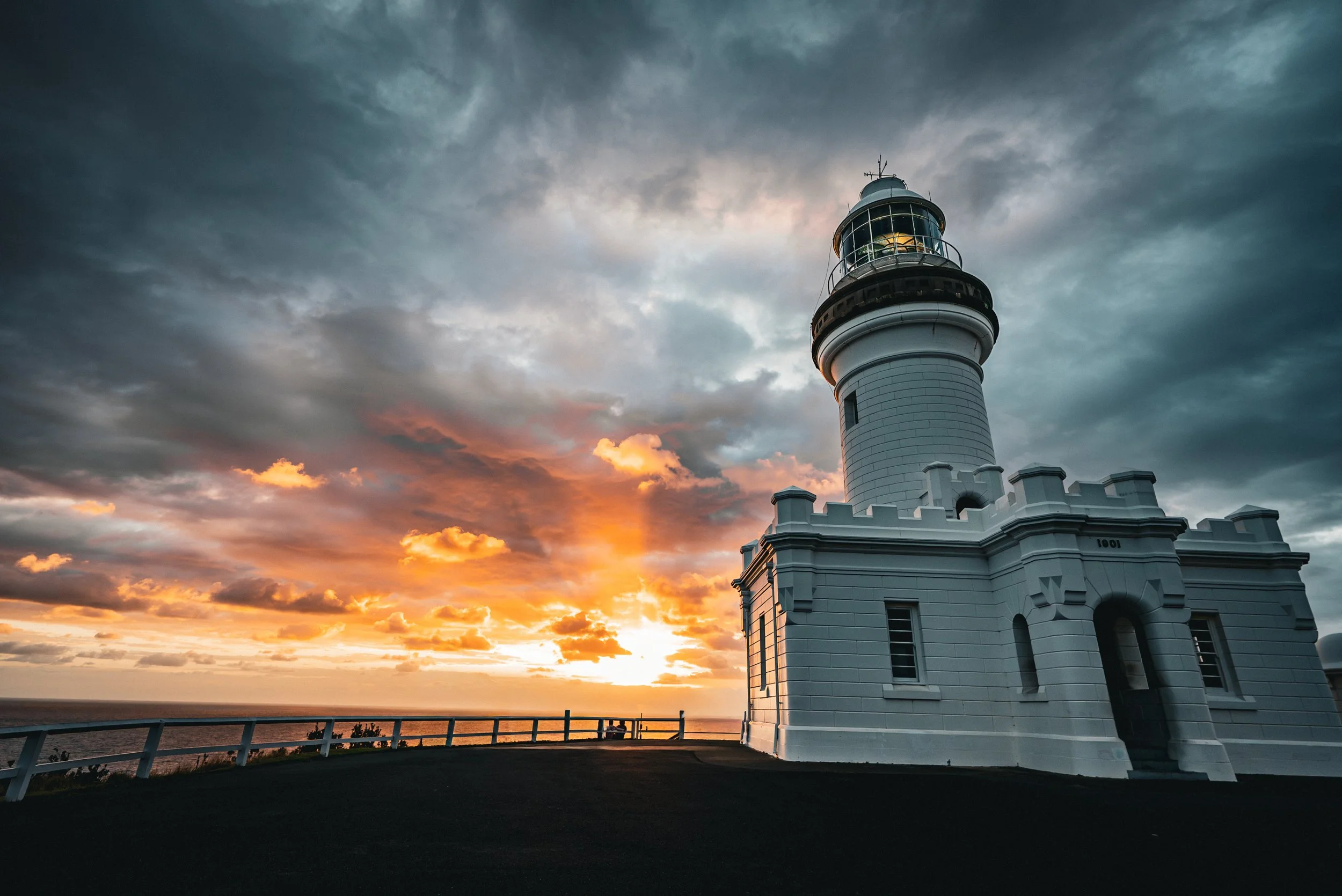 A white lighthouse with a black railing around the top stands against a stormy sky during sunset, with the ocean and a sunset in the background.