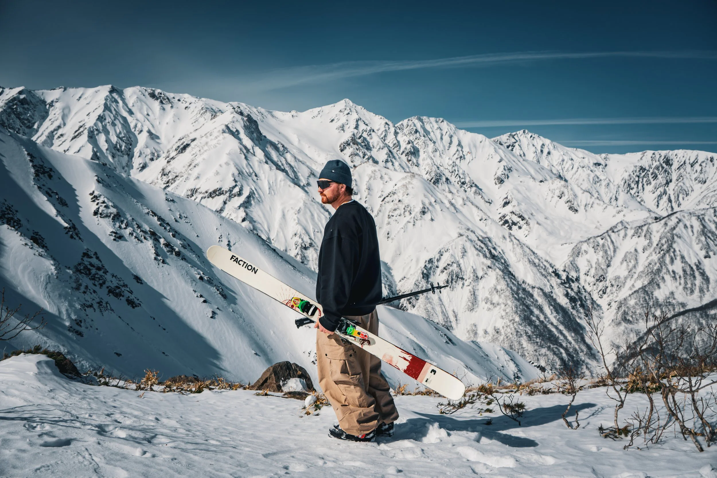 A man in winter clothing holding a ski and standing in a snowy mountain landscape with snow-covered peaks under a blue sky.