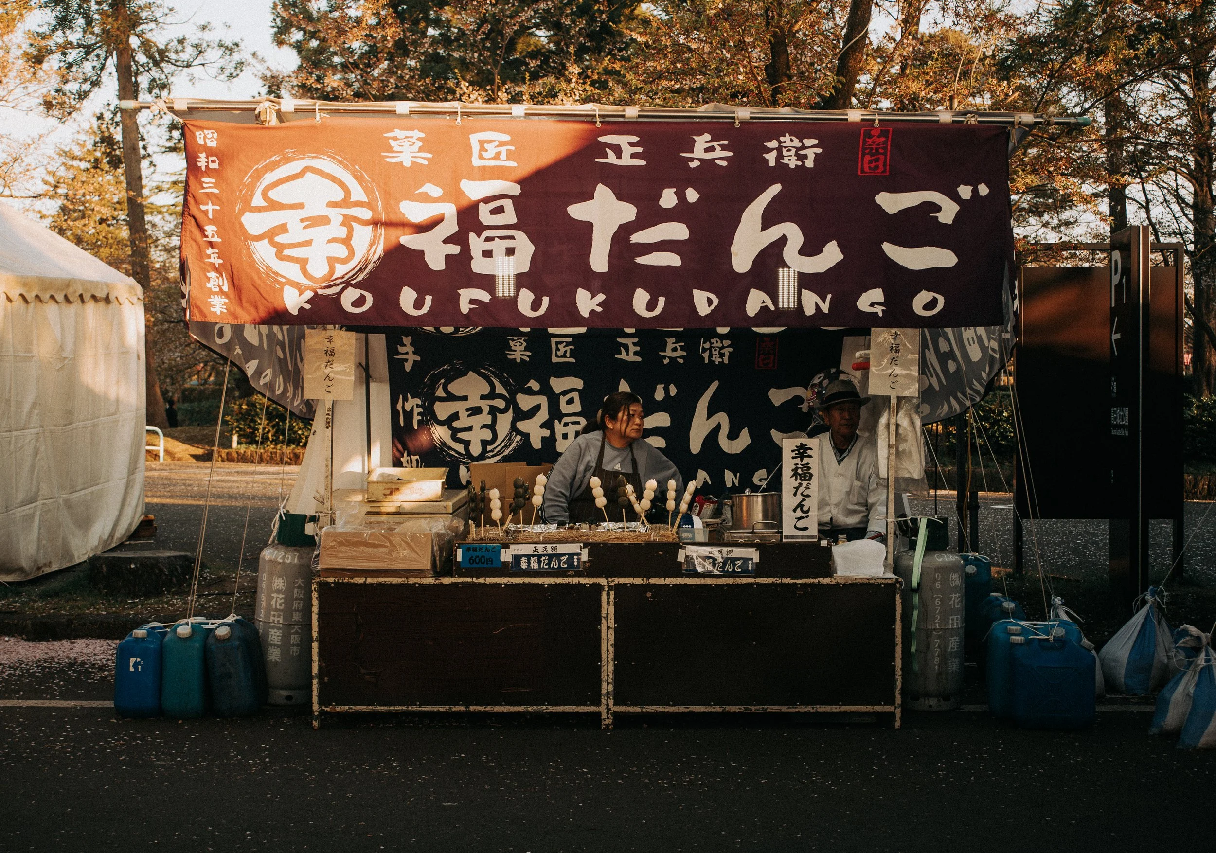 A food stand at an outdoor fair or market with a large brown and black banner displaying Japanese characters and the phrase "Koufuku Dango". Two people are working behind the counter, preparing traditional Japanese rice flour dumplings on skewers. Th