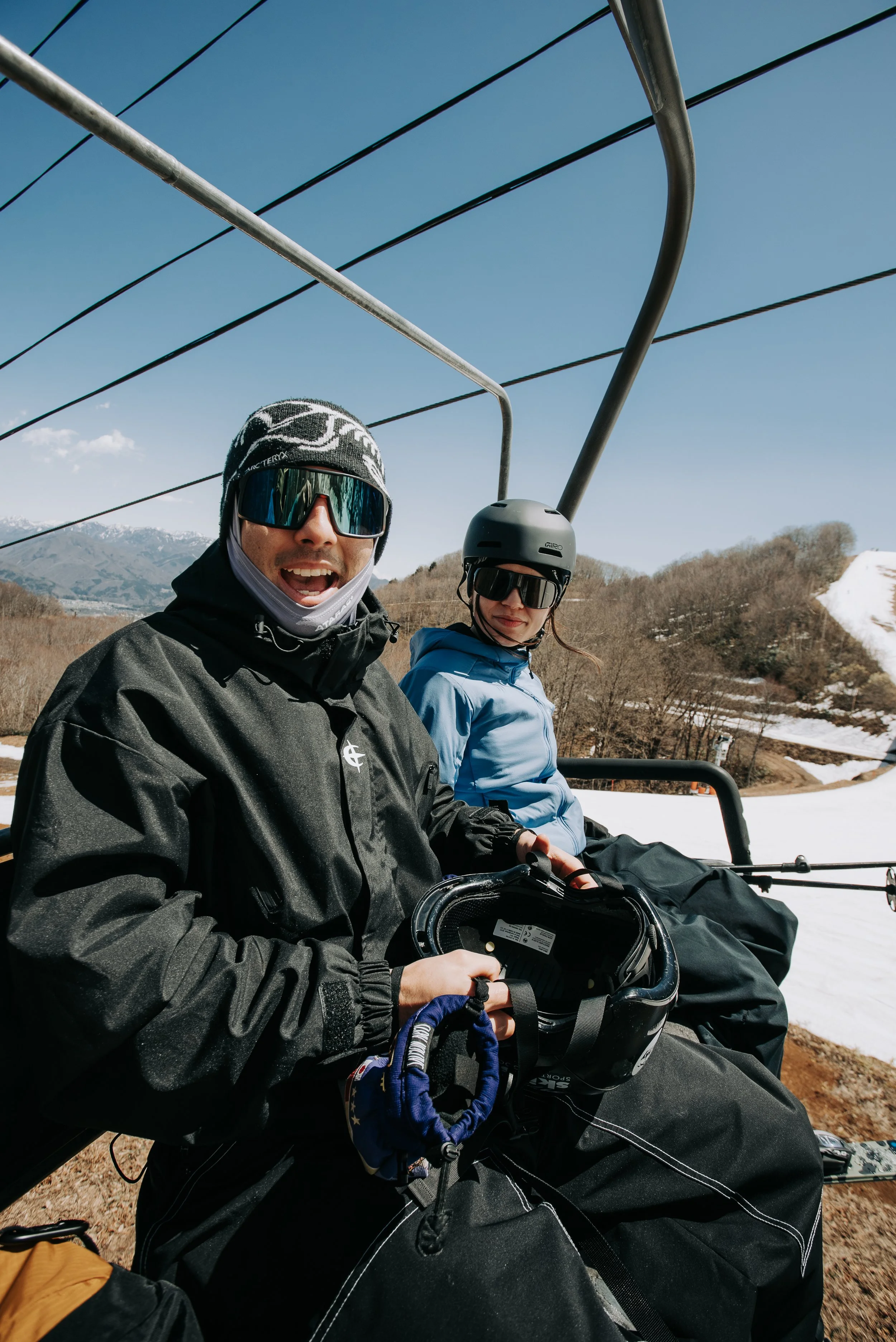 Two people riding a ski lift in a mountain area with snow patches and bare trees against a blue sky.