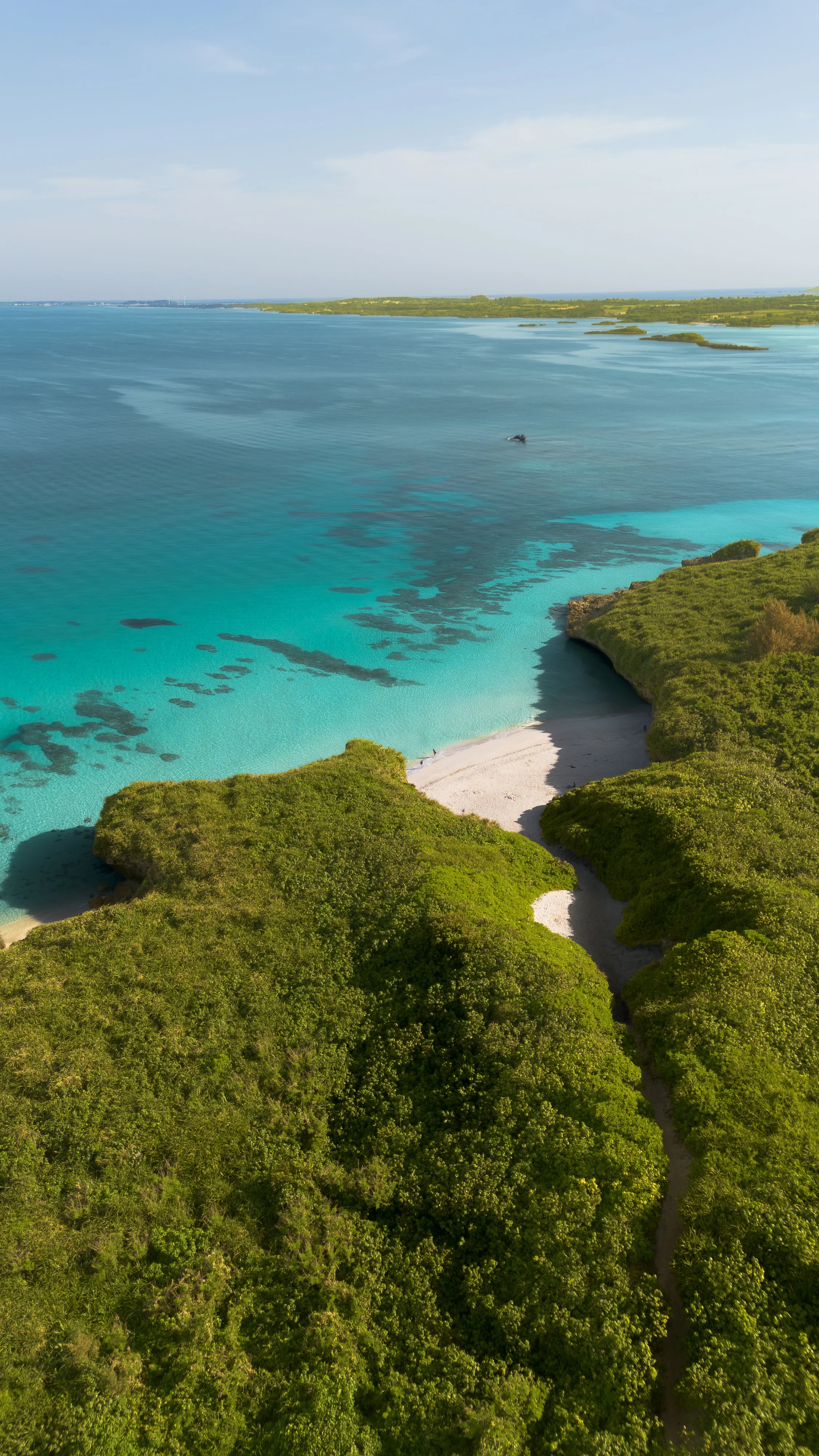 Aerial view of a tropical coastline with clear blue water, small sandy beach, lush green vegetation, and small islands in the distance under a partly cloudy sky.