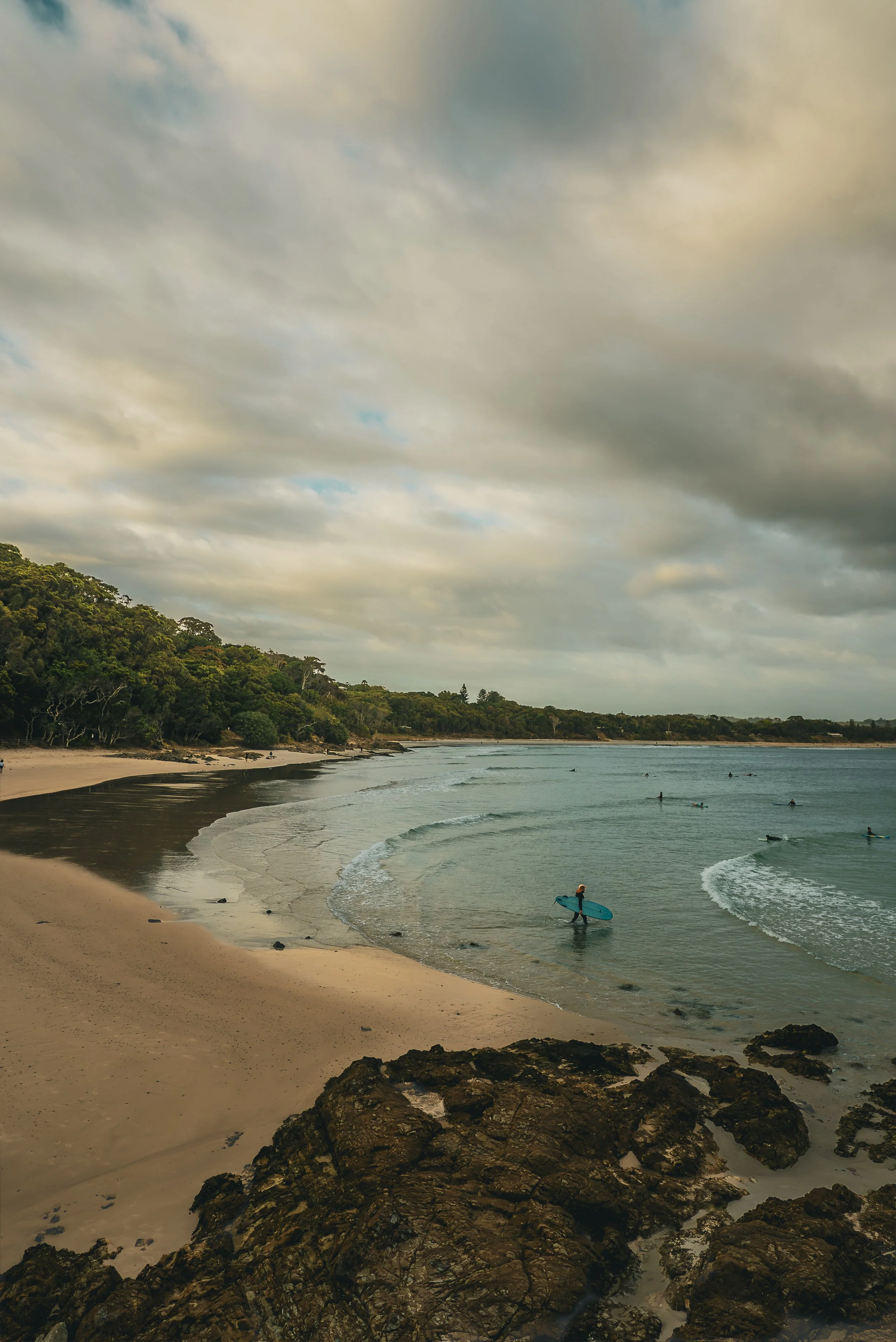 Overcast sky over a beach with surfers, rocky shoreline in the foreground, forested hillside in the background.