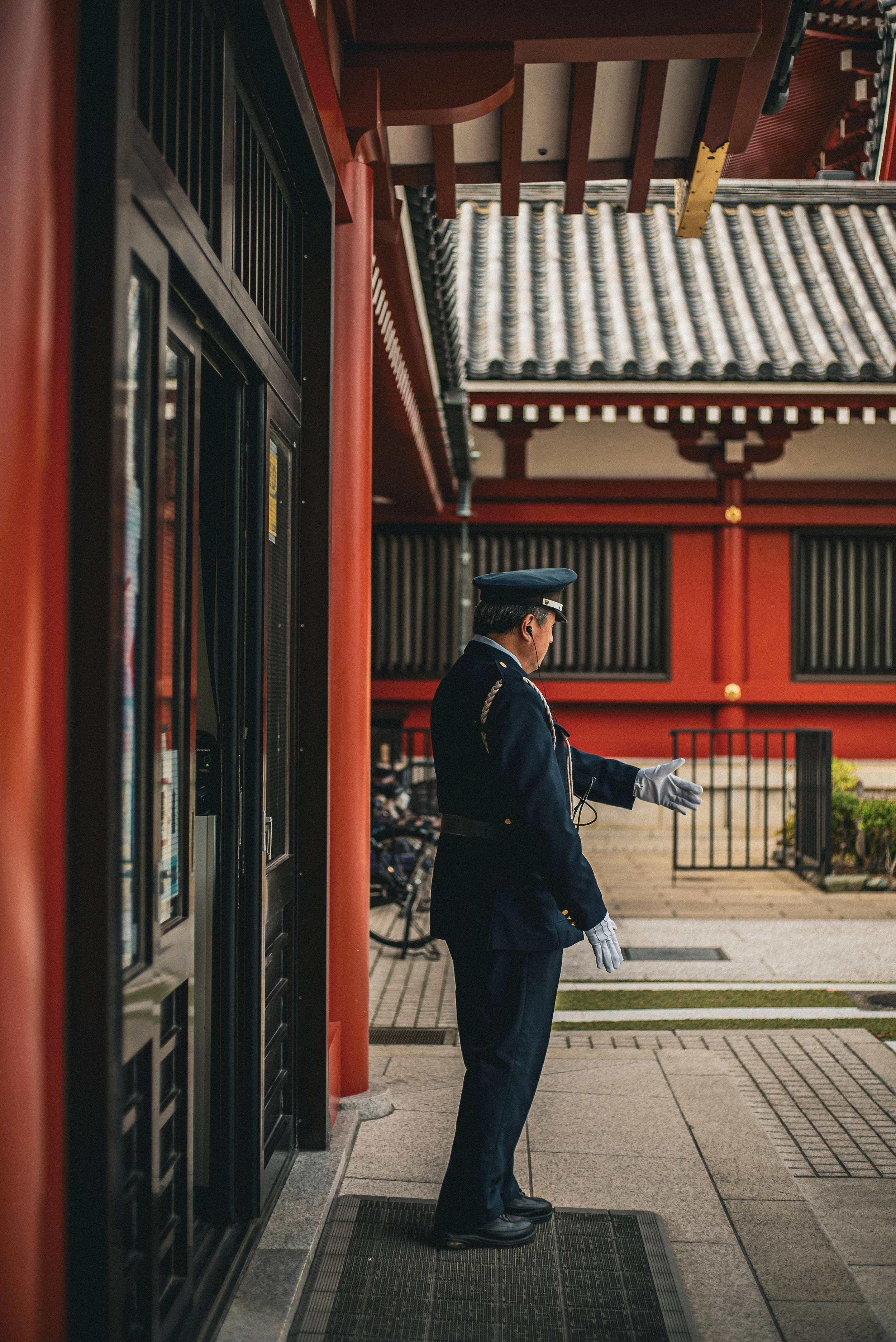 A man in a formal uniform with a hat, white gloves, and earphones standing outside a traditional Asian building with red and black decor, under a tiled roof.