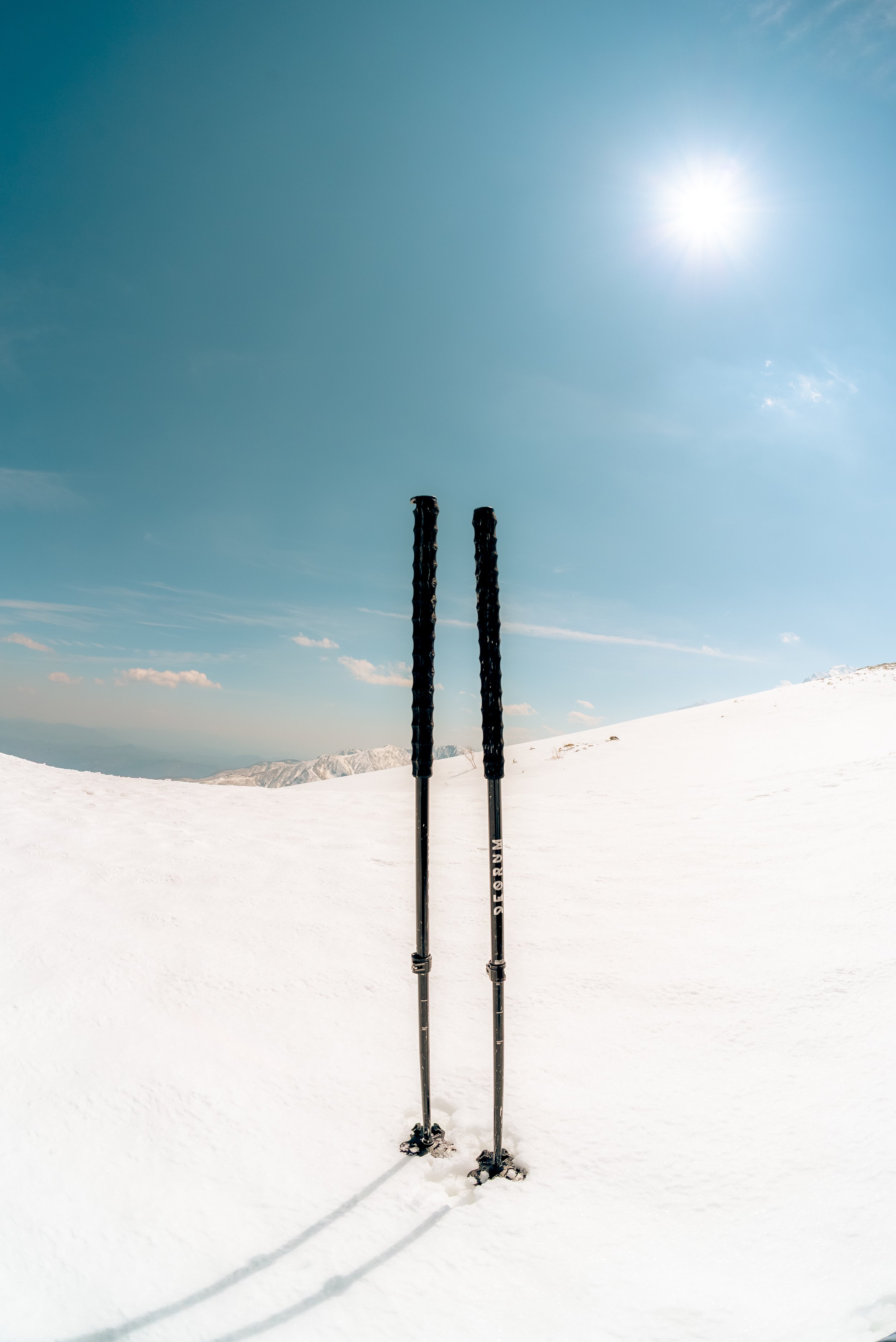 Two ski poles standing upright on snow, with a snowy mountain range in the background and a bright sun in a clear blue sky.