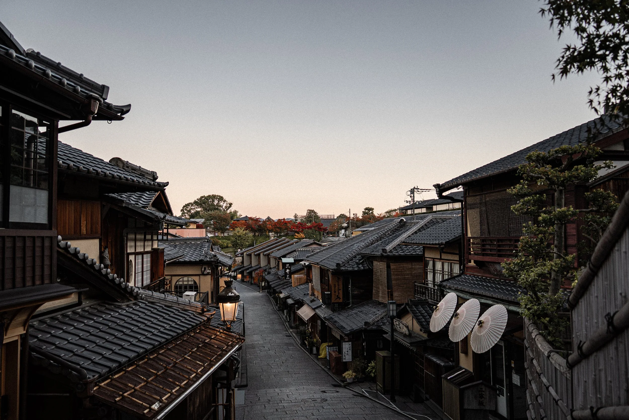 A quiet traditional Japanese street lined with wooden buildings and tiled roofs during sunset, with a sky painted in soft pastels and some trees with orange and green foliage in the background.