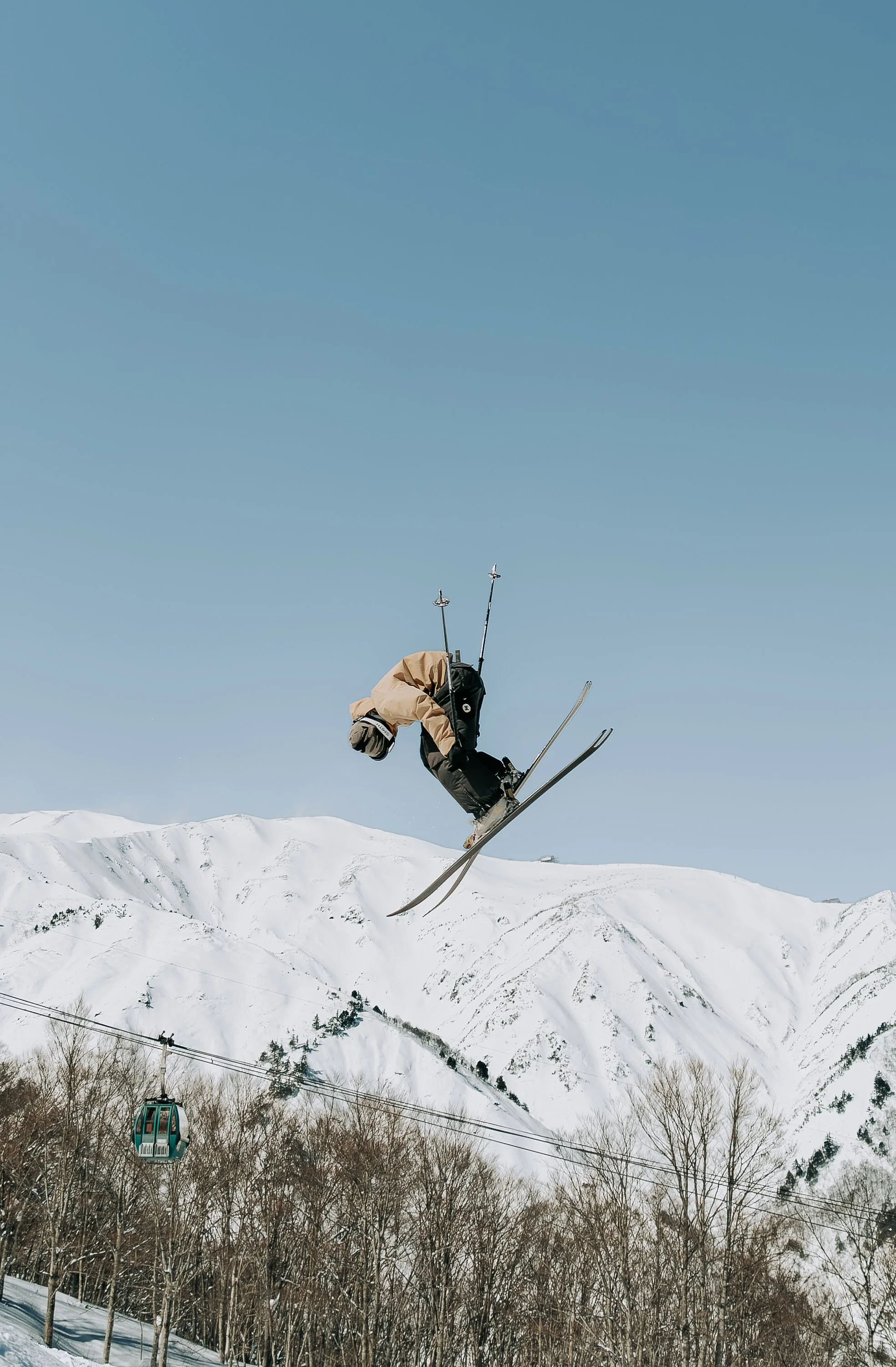 A skier in mid-air performing a jump on a snowy mountain landscape with a blue sky background and ski lift cables in the foreground.