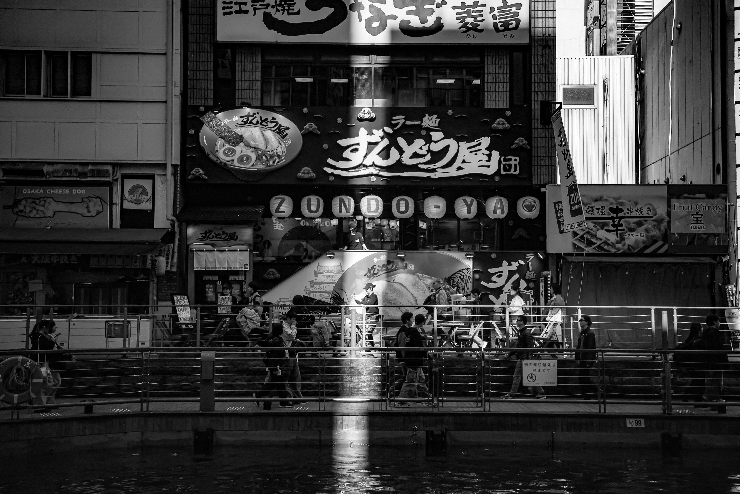 Black and white photo of a city street scene with a restaurant sign reading 'ZUNDA-YA' and people walking along a waterfront promenade with a railing.