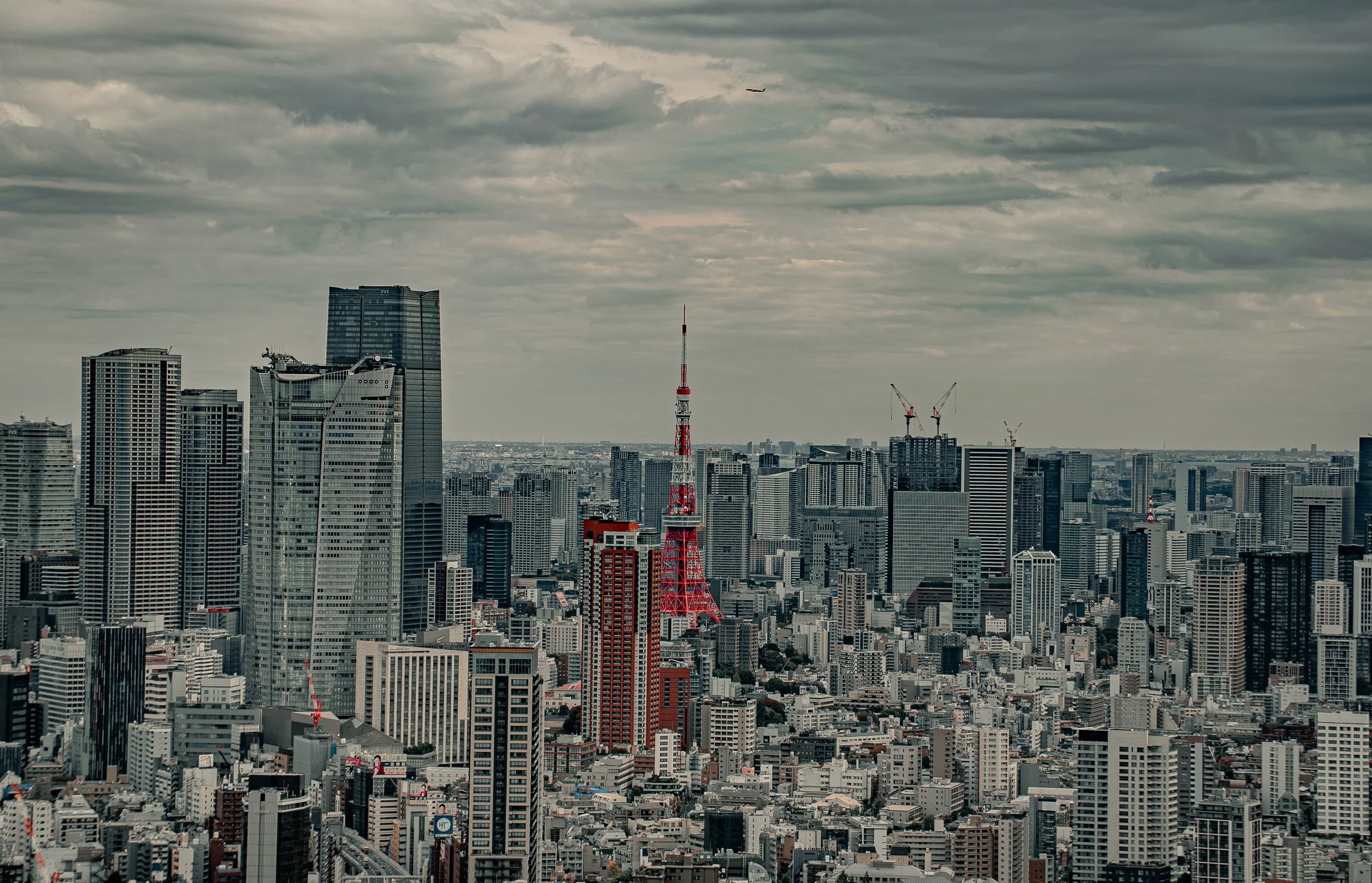 A city skyline with numerous high-rise buildings under a cloudy sky, including Tokyo Tower in the foreground and an airplane flying in the distance.