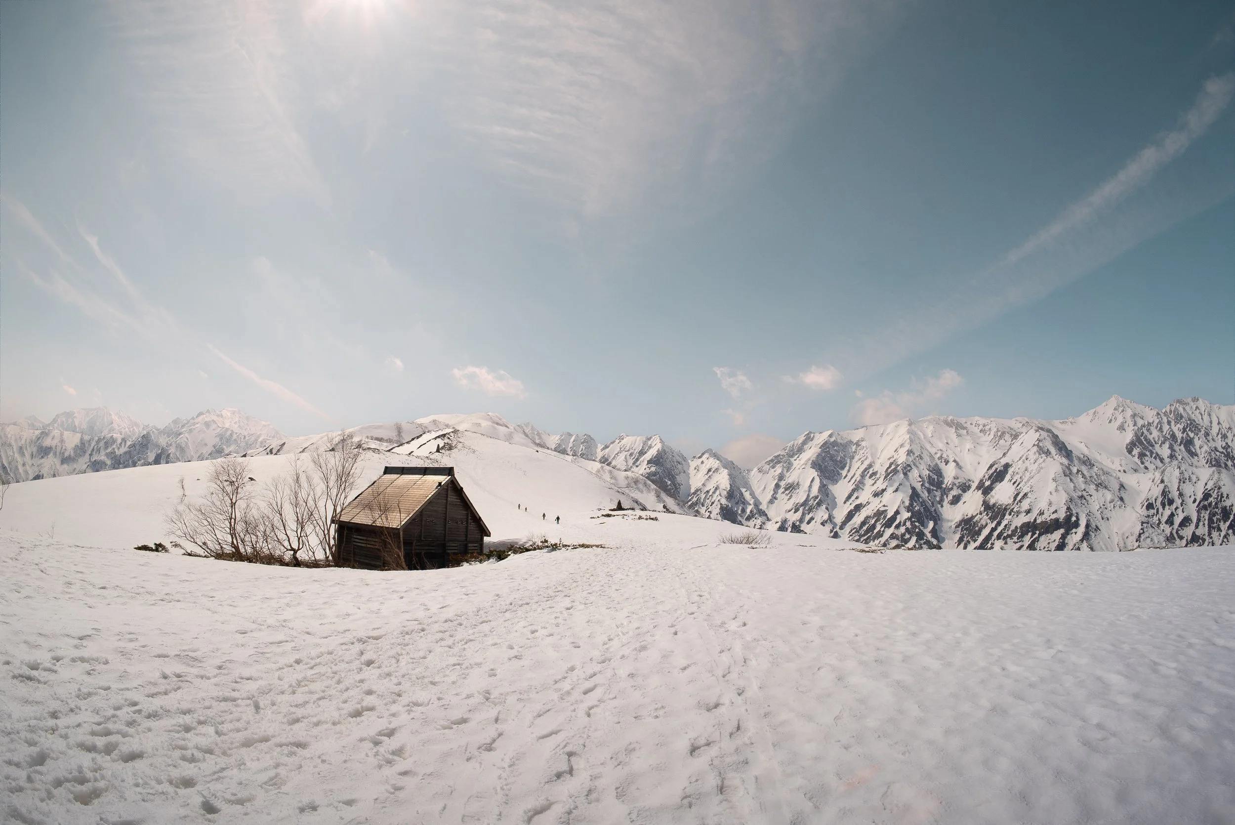 Snow-covered landscape with a small wooden cabin, leafless trees, and a background of snow-capped mountains under a partly cloudy sky.