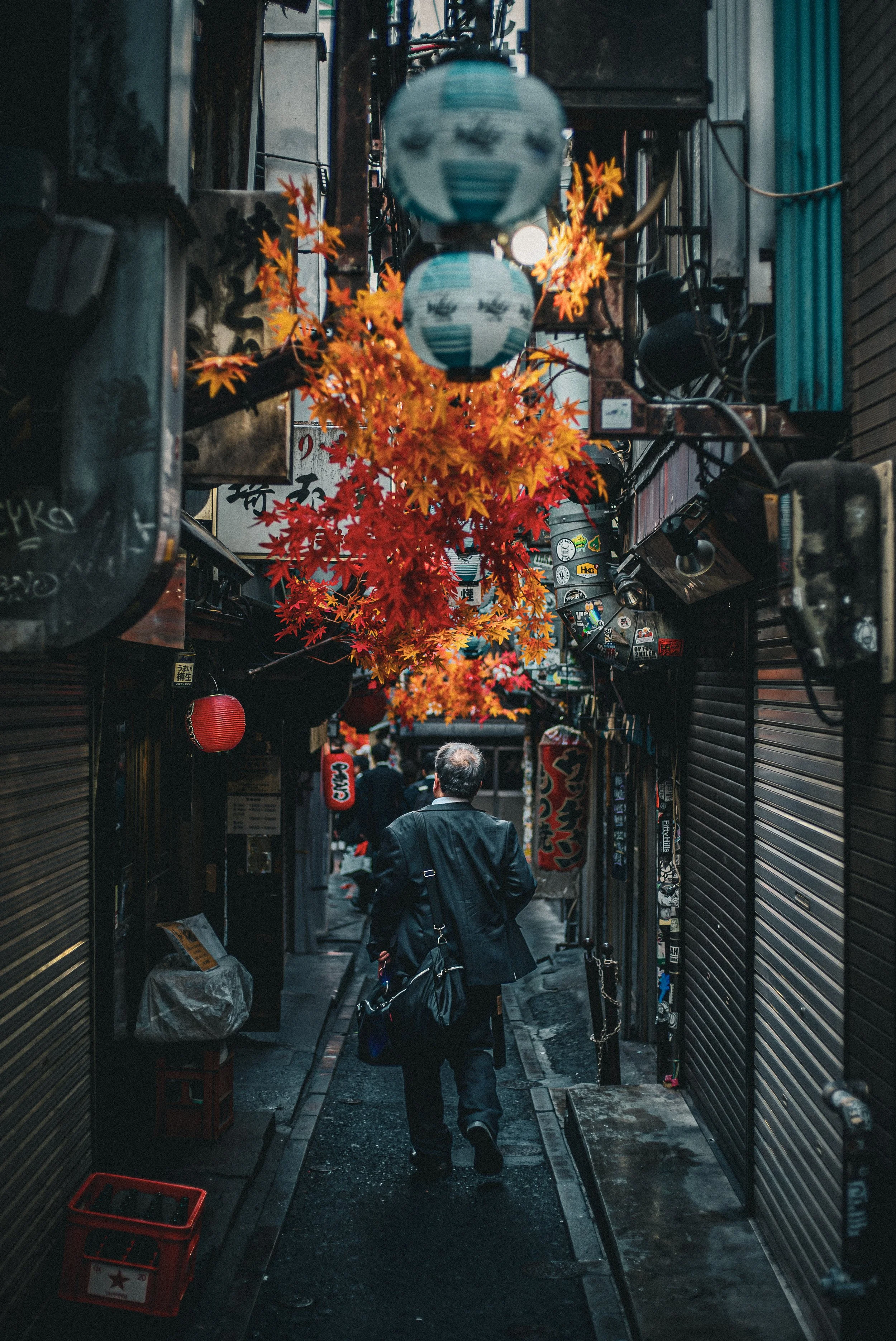 A narrow alleyway with shops on both sides, decorated with orange and red maple leaves hanging overhead, and red lanterns and signage on the walls. A man in a black jacket with a bag is walking away from the camera.