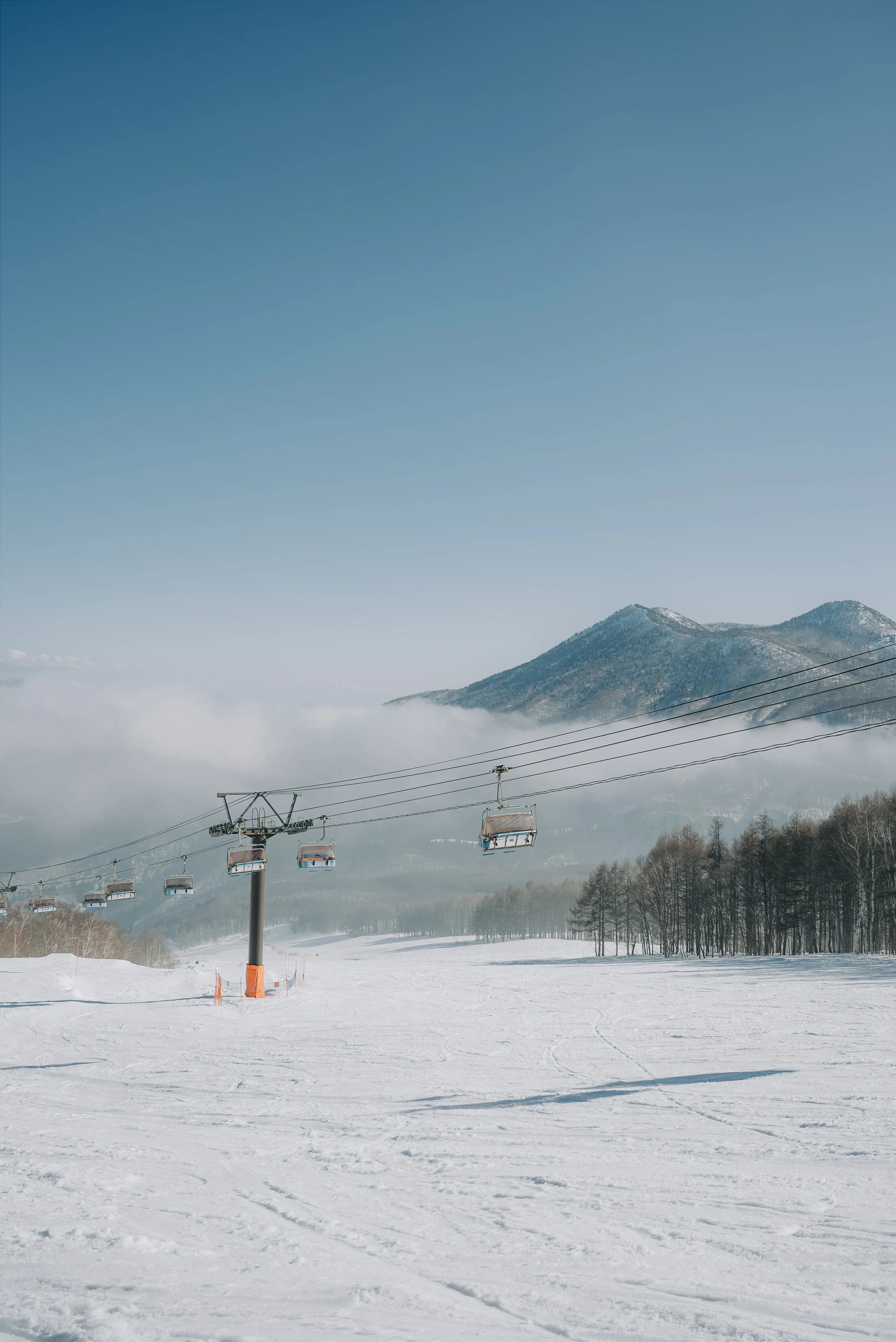 Snow-covered ski resort with ski lift chairs moving up a mountain under a clear blue sky, with snow-covered trees and distant mountain peaks in the background.