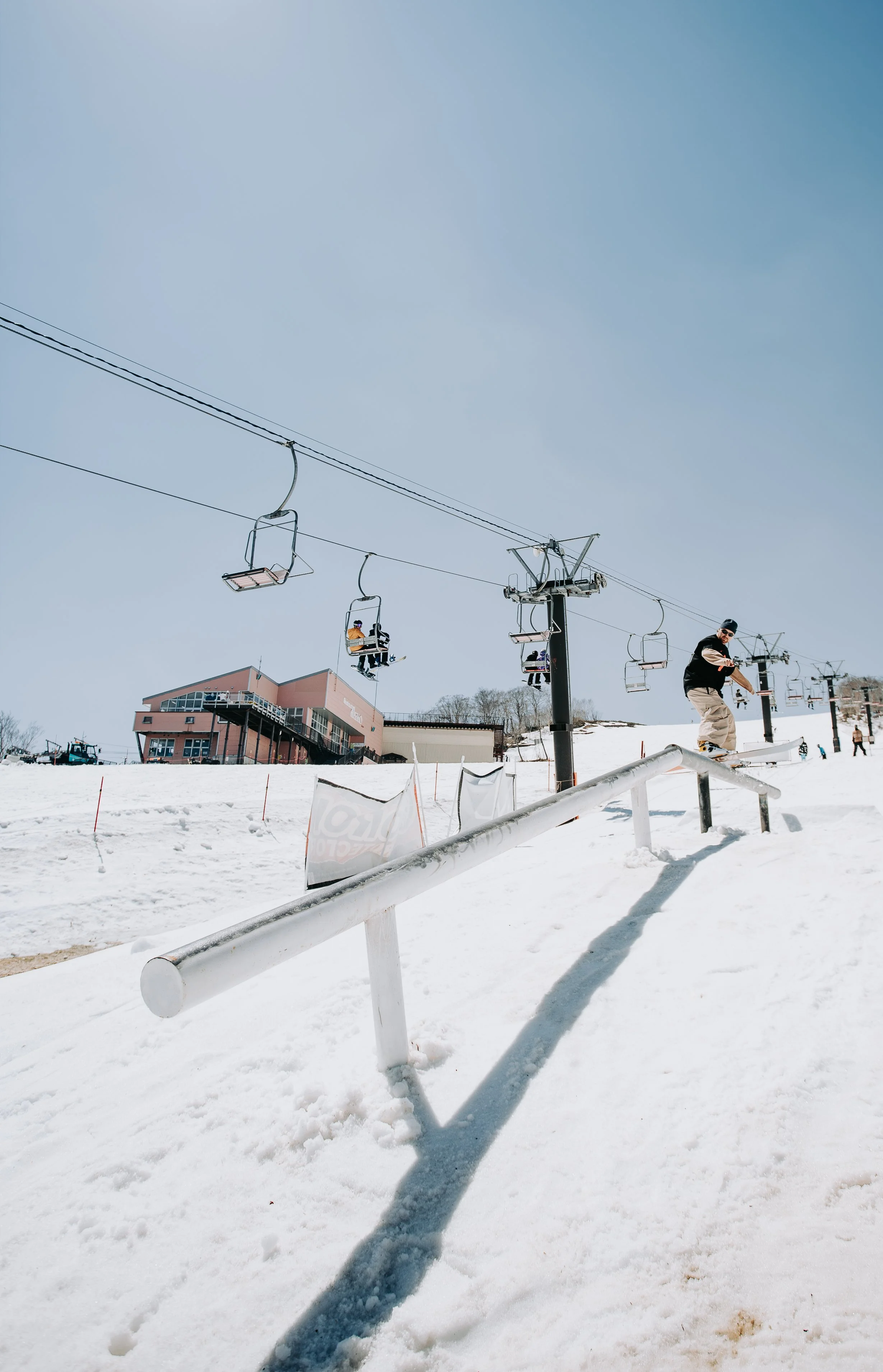 A snowy ski resort with a ski lift and a skier performing a trick on a rail. Buildings and trees are in the background under a clear blue sky.