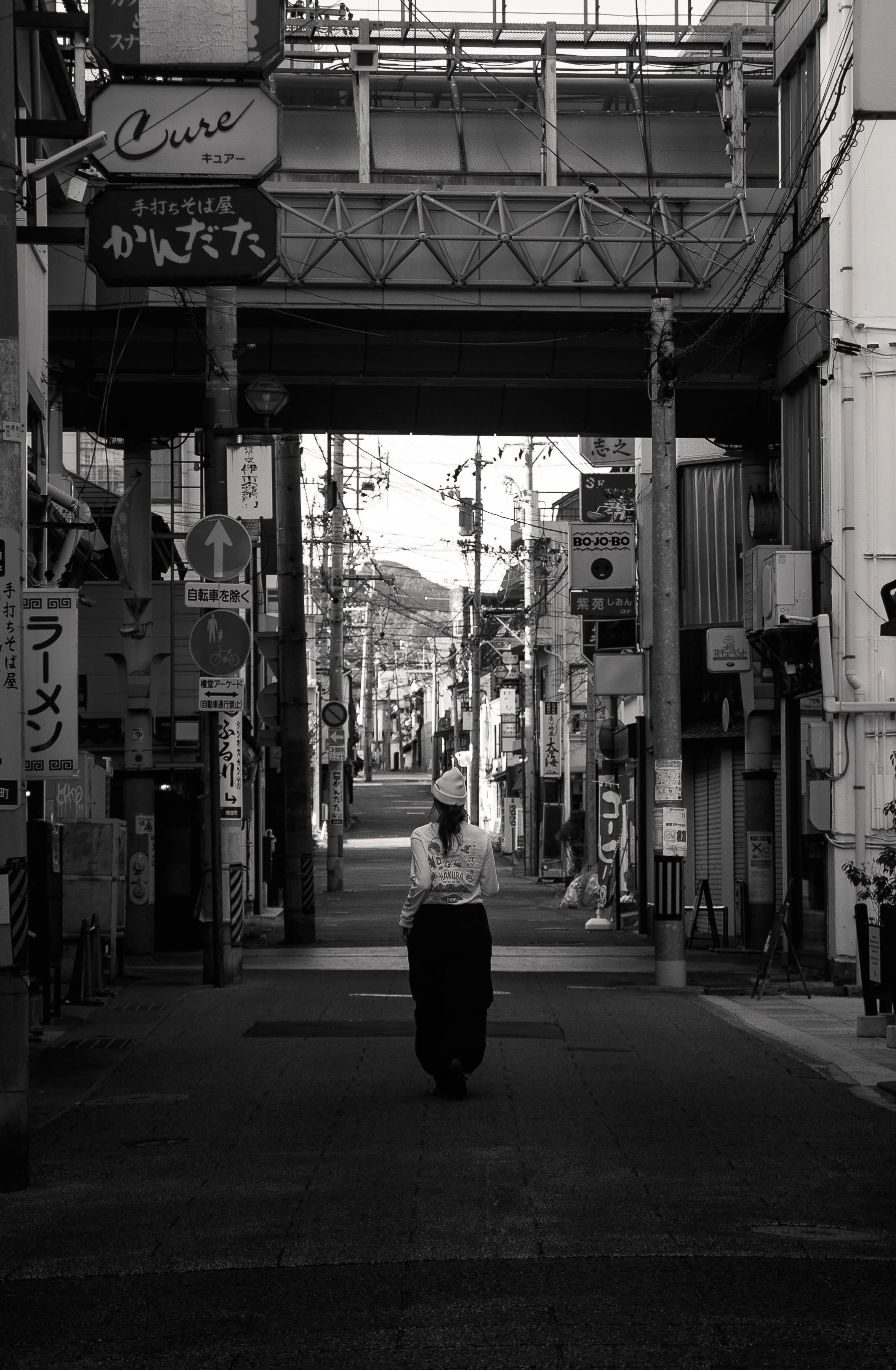 A black and white photo of a woman walking down a narrow street in Japan, surrounded by signs and wires, with a bridge overhead.