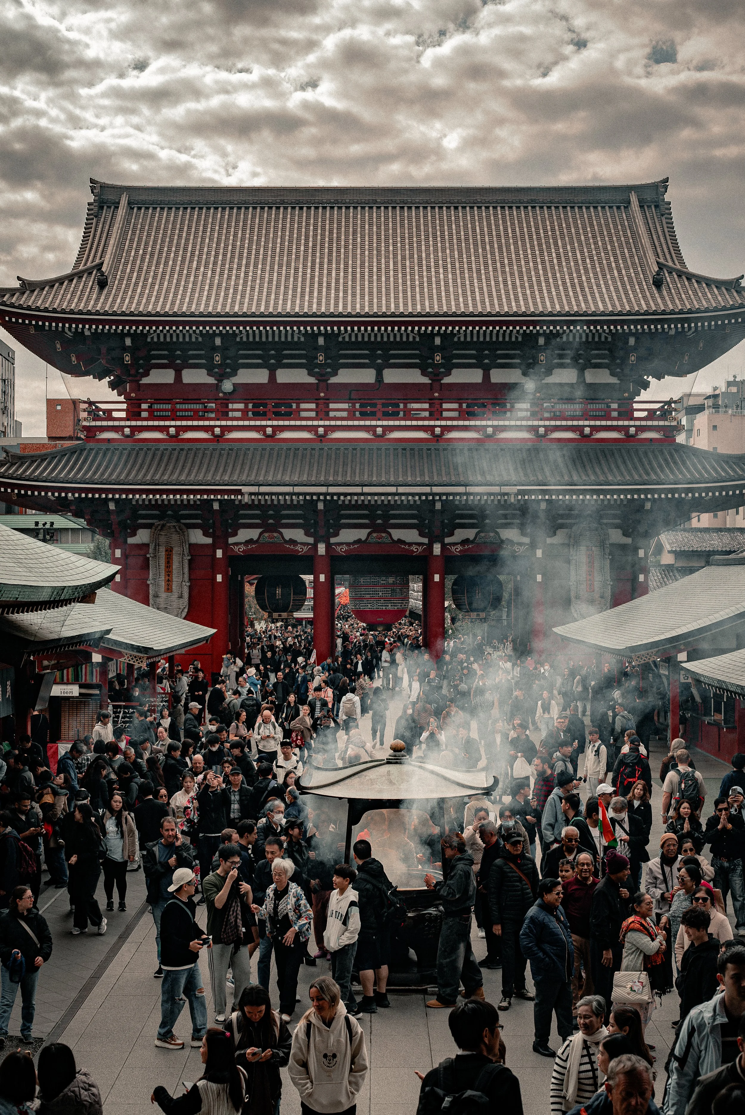 Crowd of people gathered in front of a traditional Japanese temple with a large, intricately designed roof, and a smoking incense burner in the foreground.