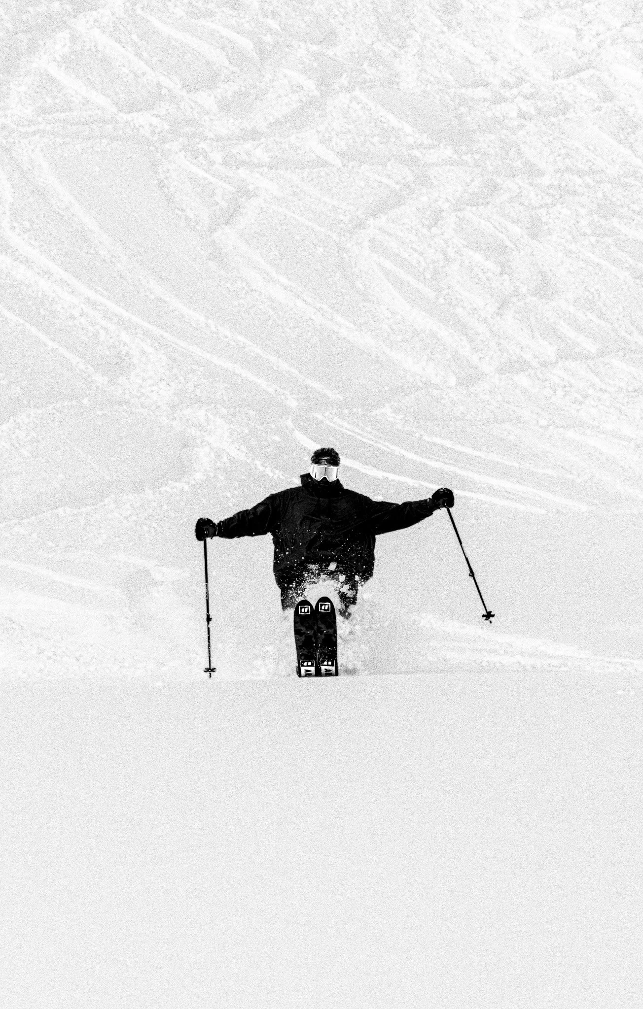 A skier wearing black gear and goggles skiing down a snowy slope with a mountain in the background.