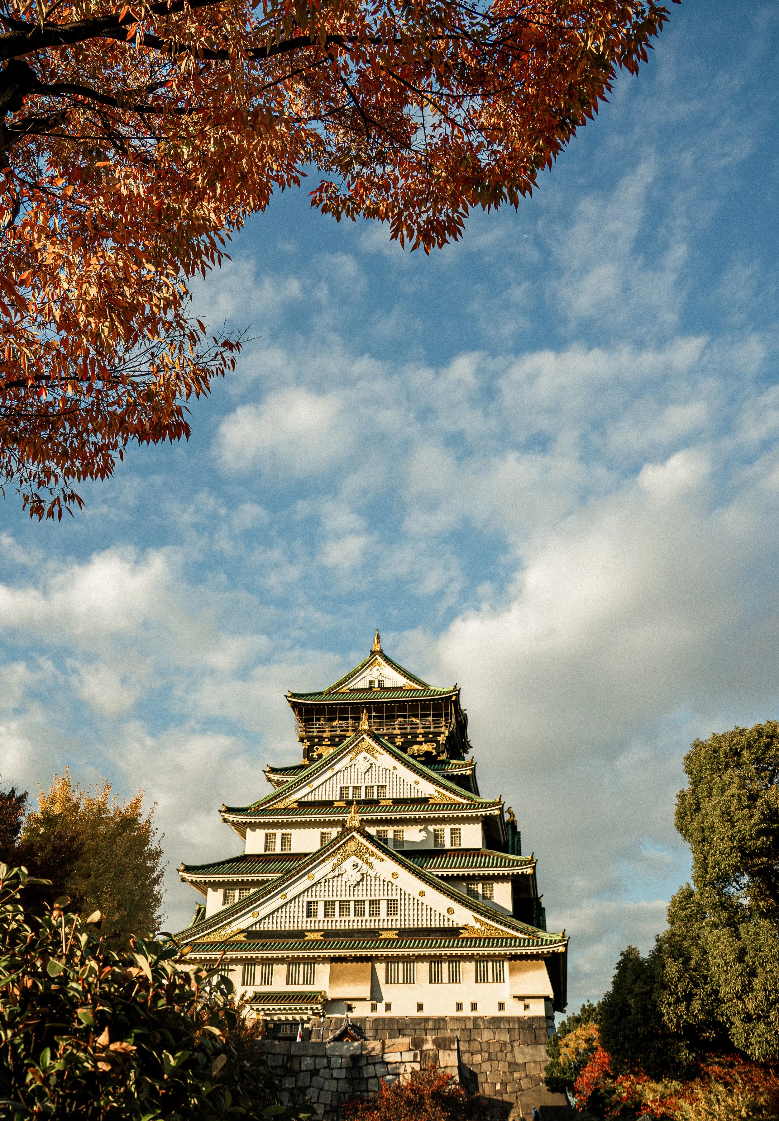 A traditional Japanese castle with multiple tiers and green roofs, surrounded by trees with autumn-colored leaves, under a partly cloudy sky.