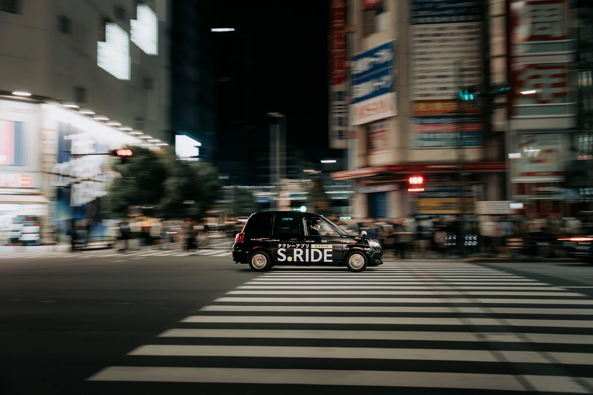 A black taxi driving through a city street at night, with illuminated billboards and buildings in the background, and a crosswalk in the foreground.