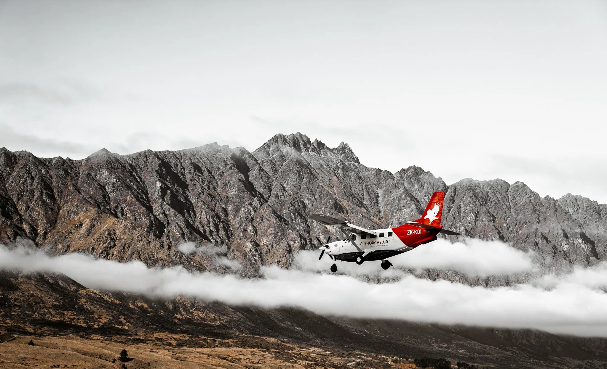 A small aircraft flying low over a mountainous landscape with rugged peaks and clouds in the background.