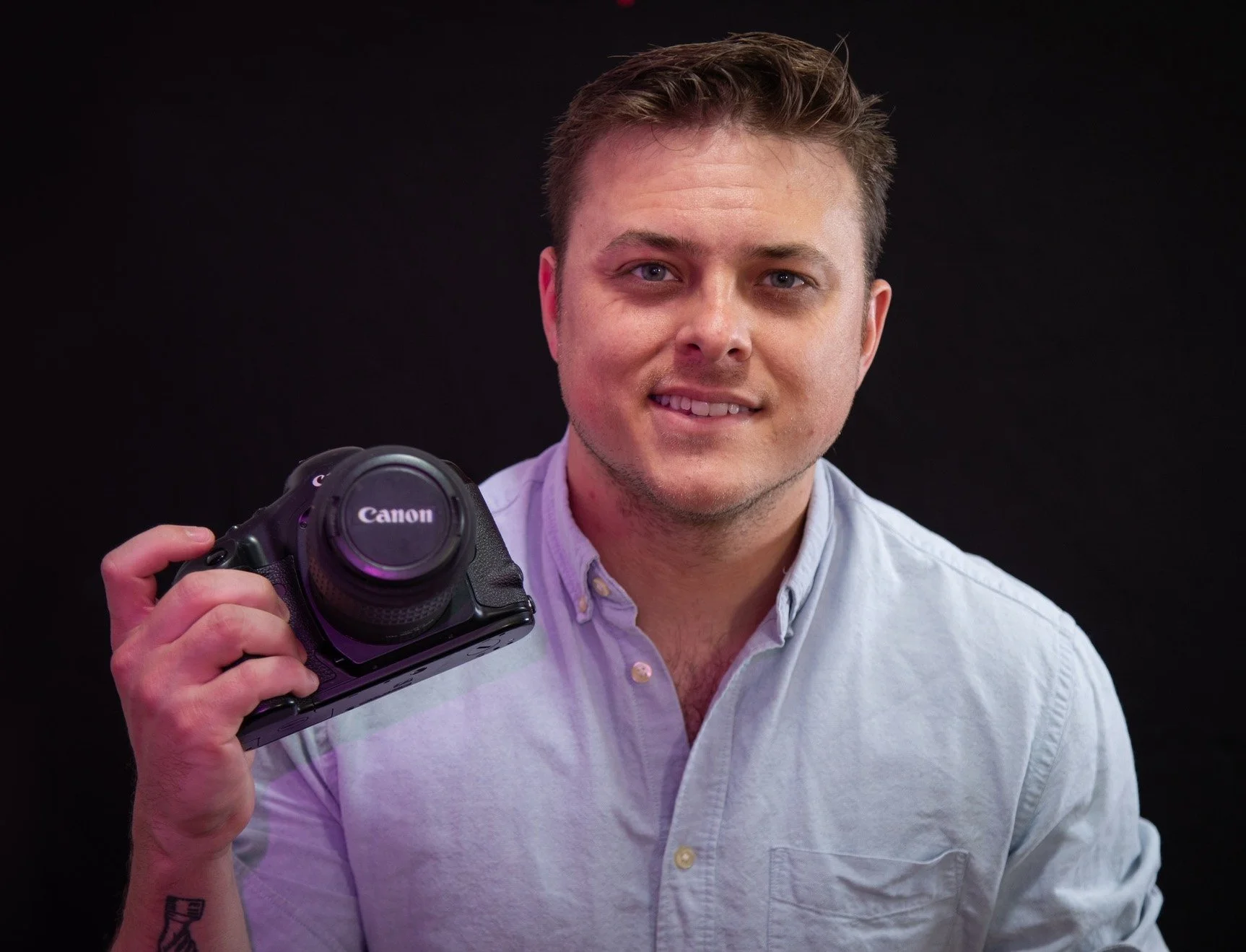 A young man with light brown hair, blue eyes, and light skin holding a Canon camera in his right hand against a black background. He is wearing a light-colored button-up shirt and smiling slightly.