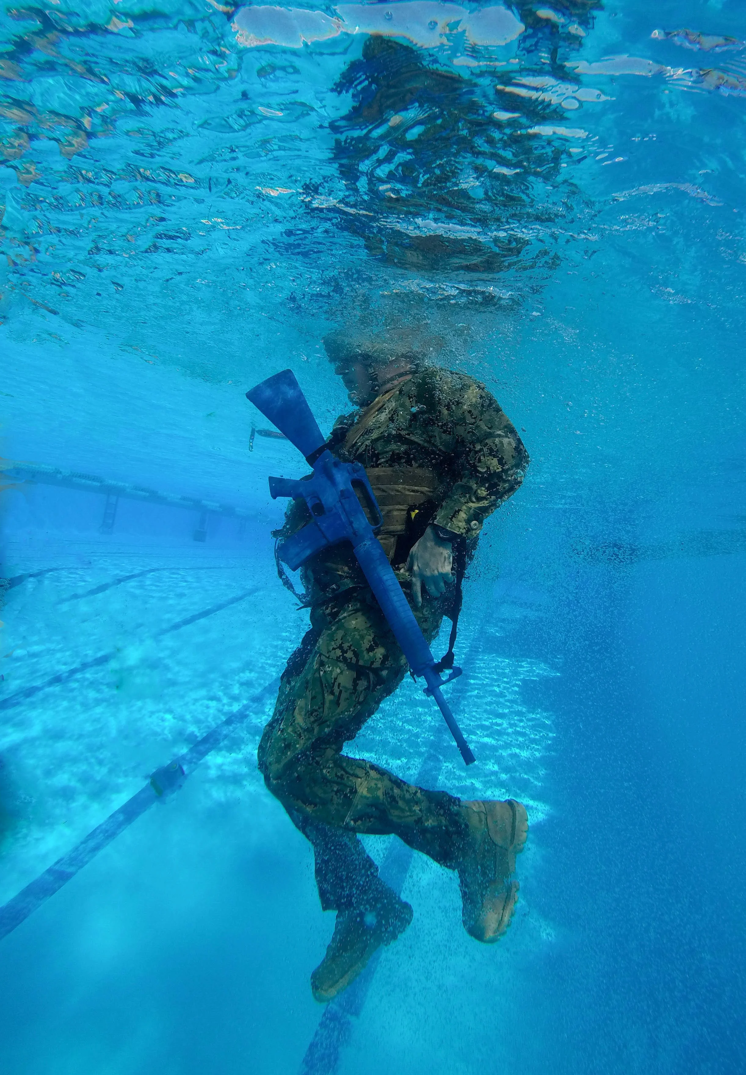A soldier wearing camouflage uniform and protective gear underwater, holding a large weapon, with the water causing bubbles around him.