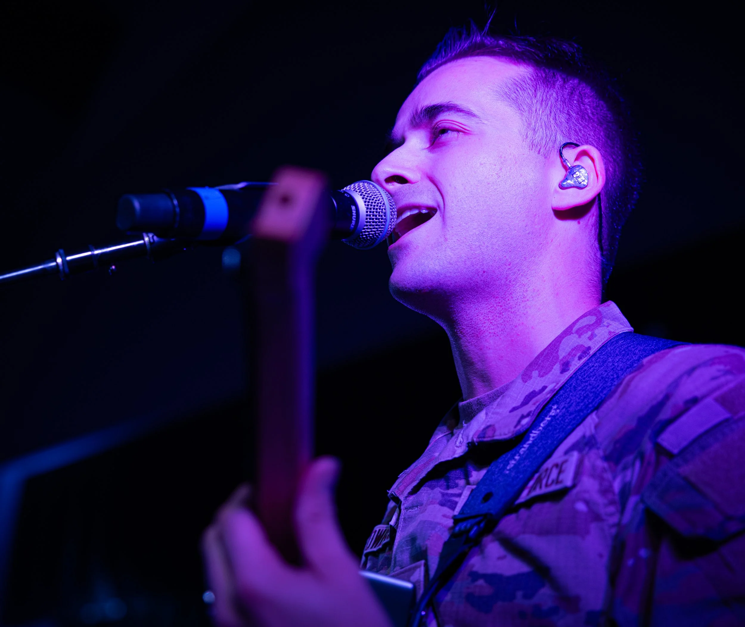 A young man singing or speaking into a microphone on stage, wearing in-ear monitors, with purple stage lighting highlighting his face and hair.