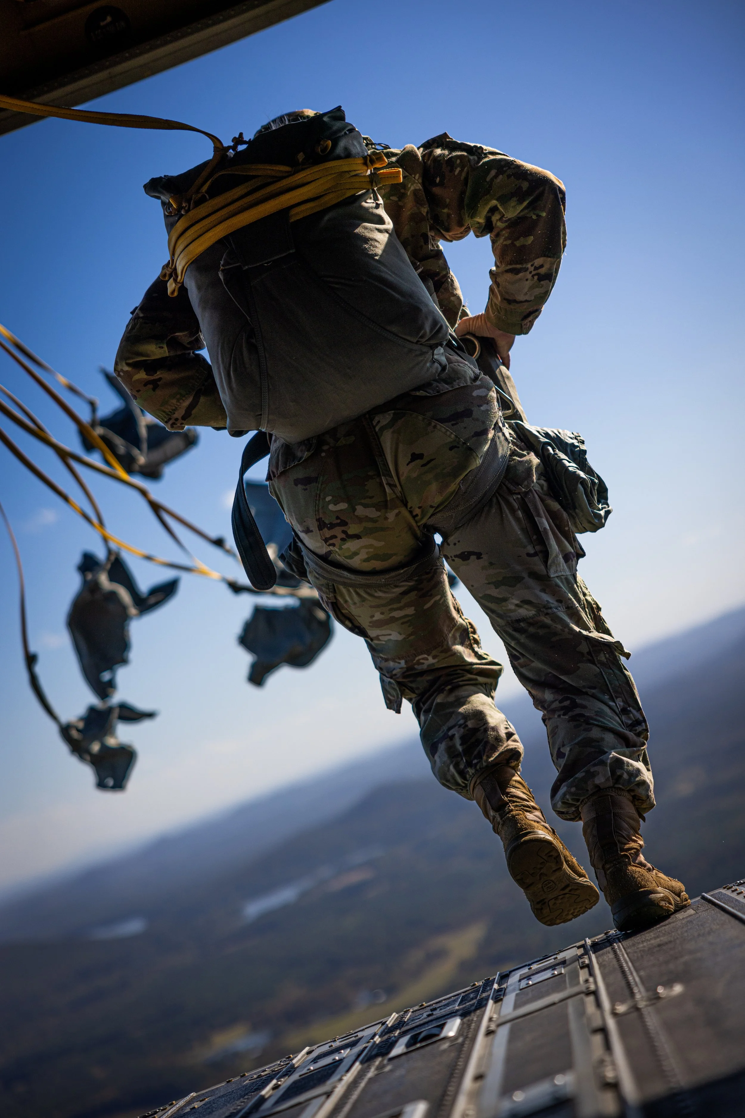 A soldier in camouflage uniform is rappelling down the side of a military aircraft, holding onto a rope with both hands. The sky is clear with a blue background, and the photo is taken from below the soldier, capturing the aircraft's side and the gro