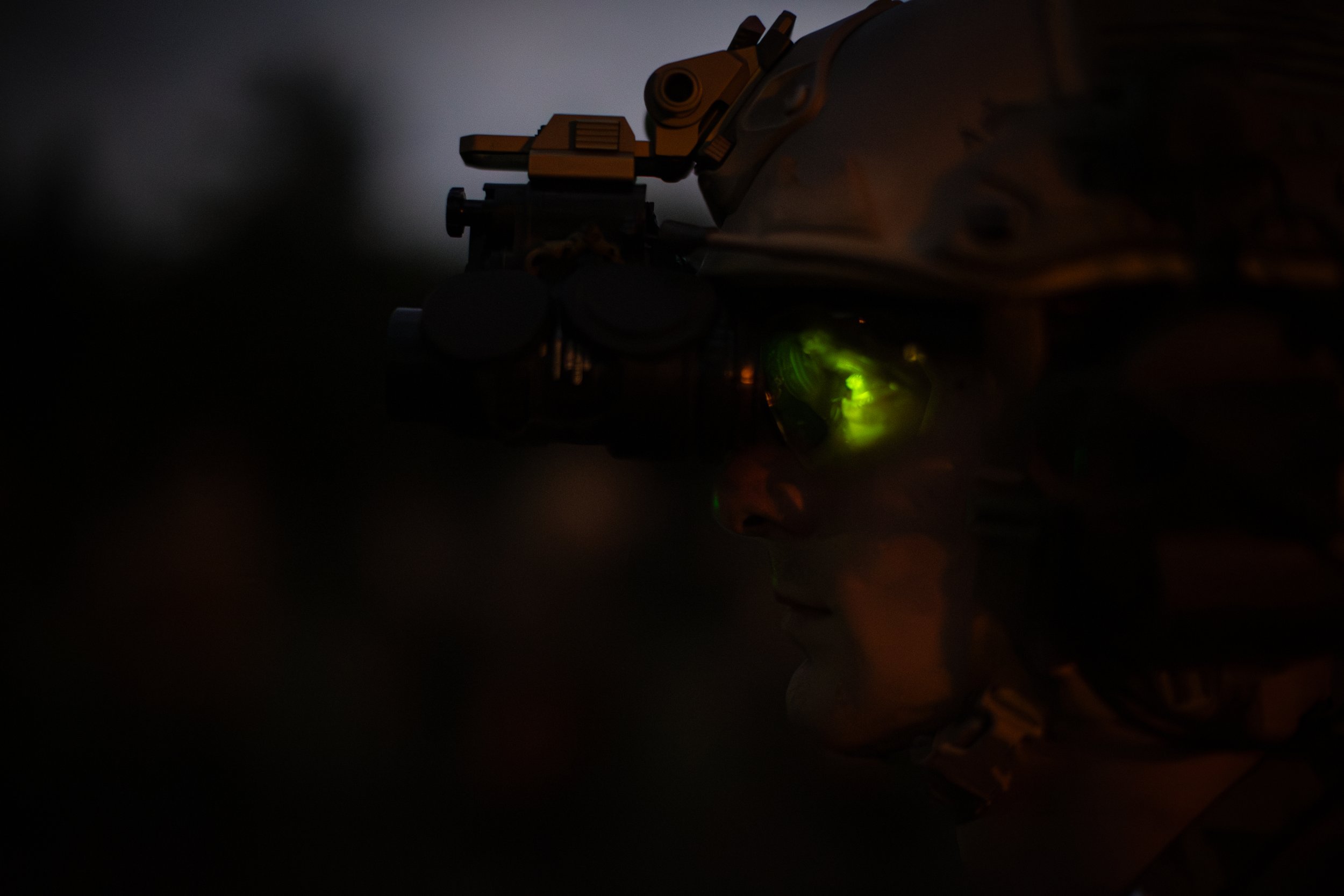 A dark image showing a person wearing a camouflage jacket aiming a rifle in a low-light environment, with a green glow from the firearm's sight.