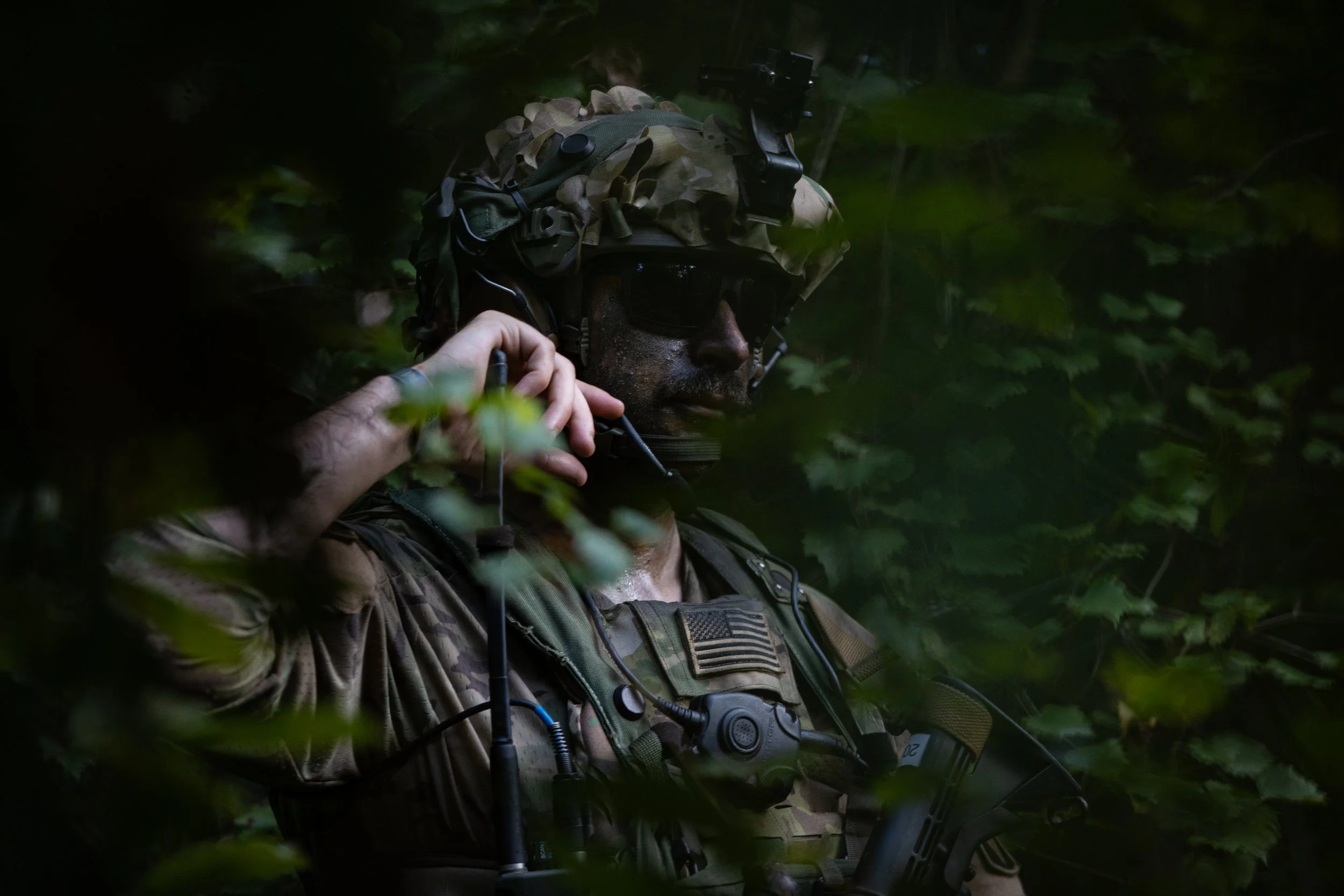 A soldier in camouflage gear, wearing a helmet and sunglasses, holds a communication device near his ear while riding a motorcycle through a green, wooded area.