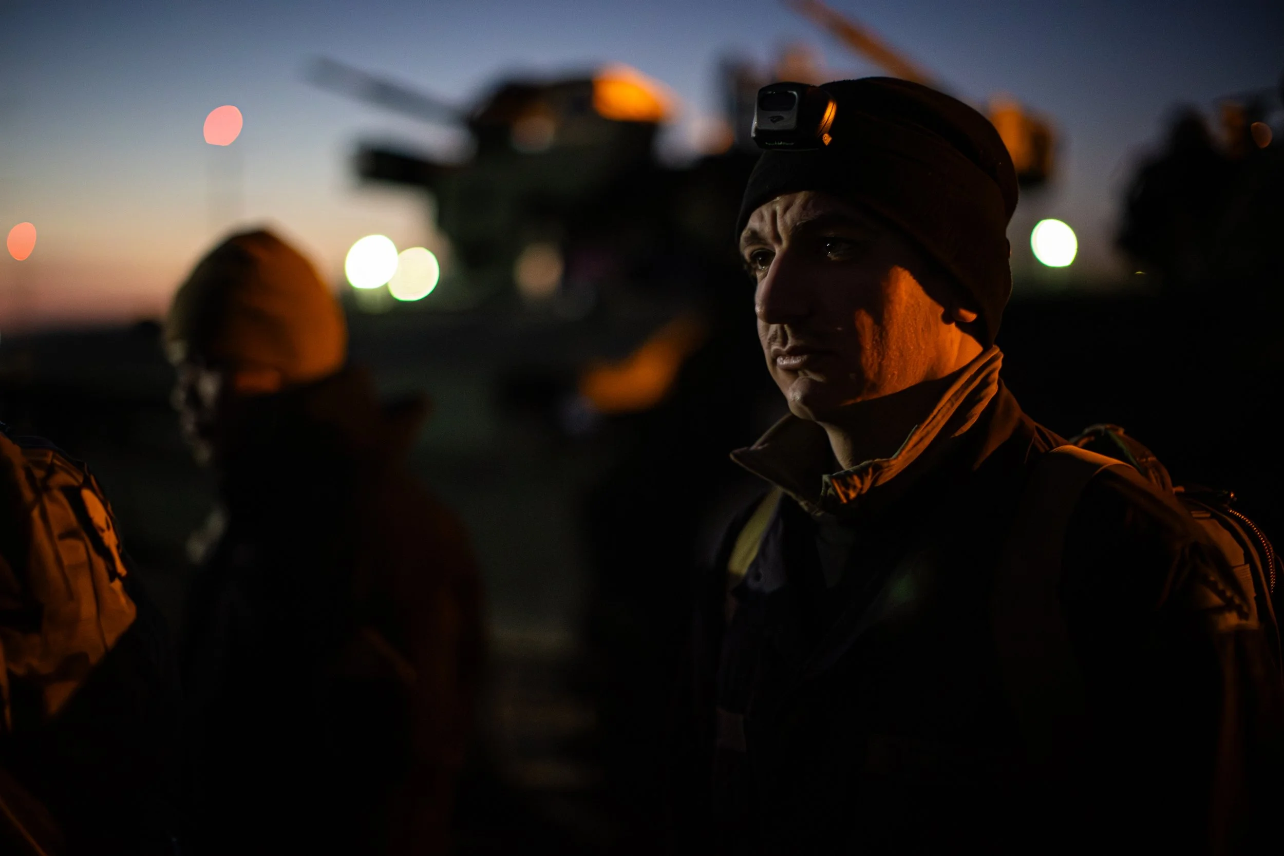 A person wearing a beanie and headlamp stands at dusk with other individuals, with a helicopter and colorful lights in the background.