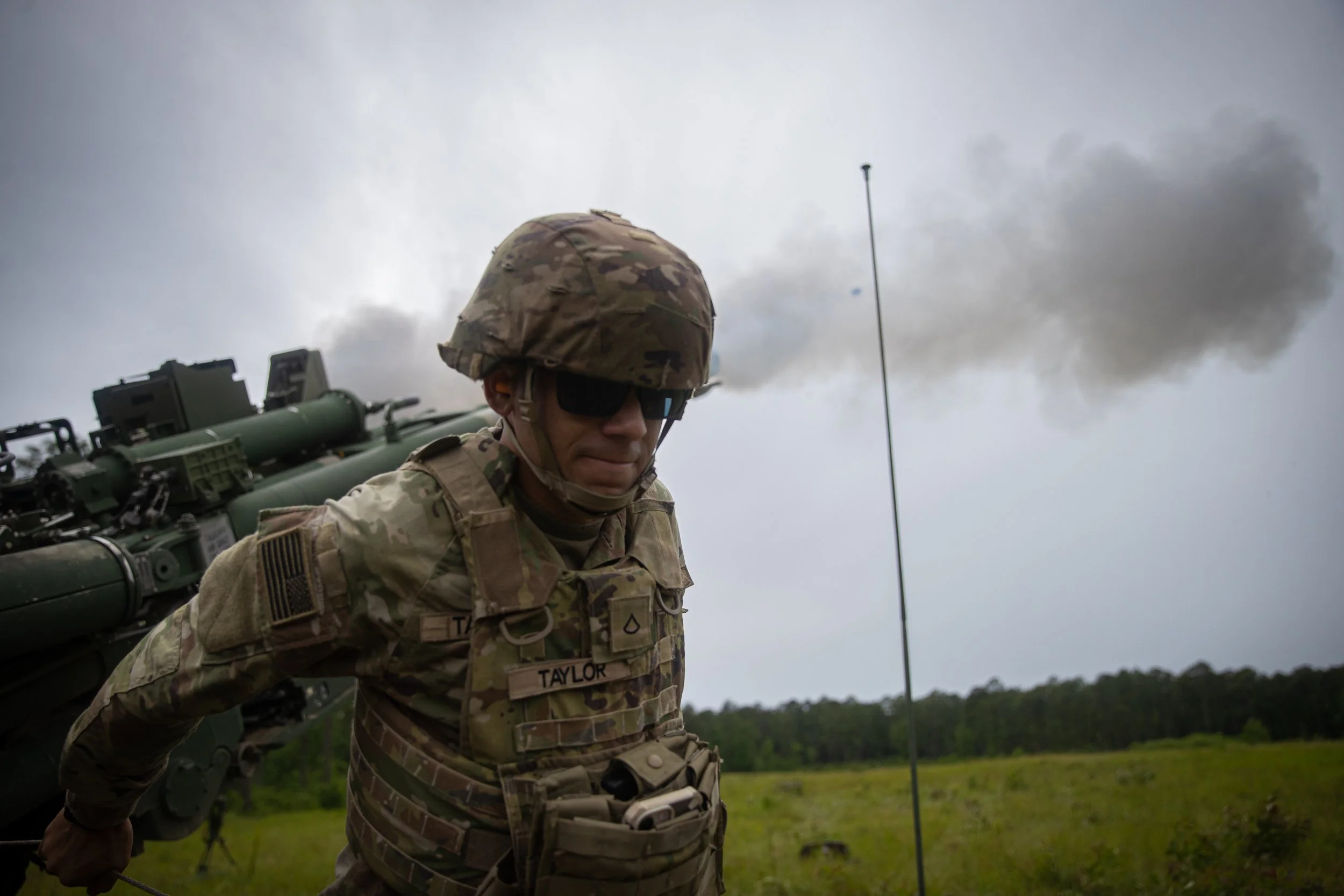 A soldier in camouflage uniform and helmet with sunglasses, standing outside under cloudy sky, with military equipment on his back.