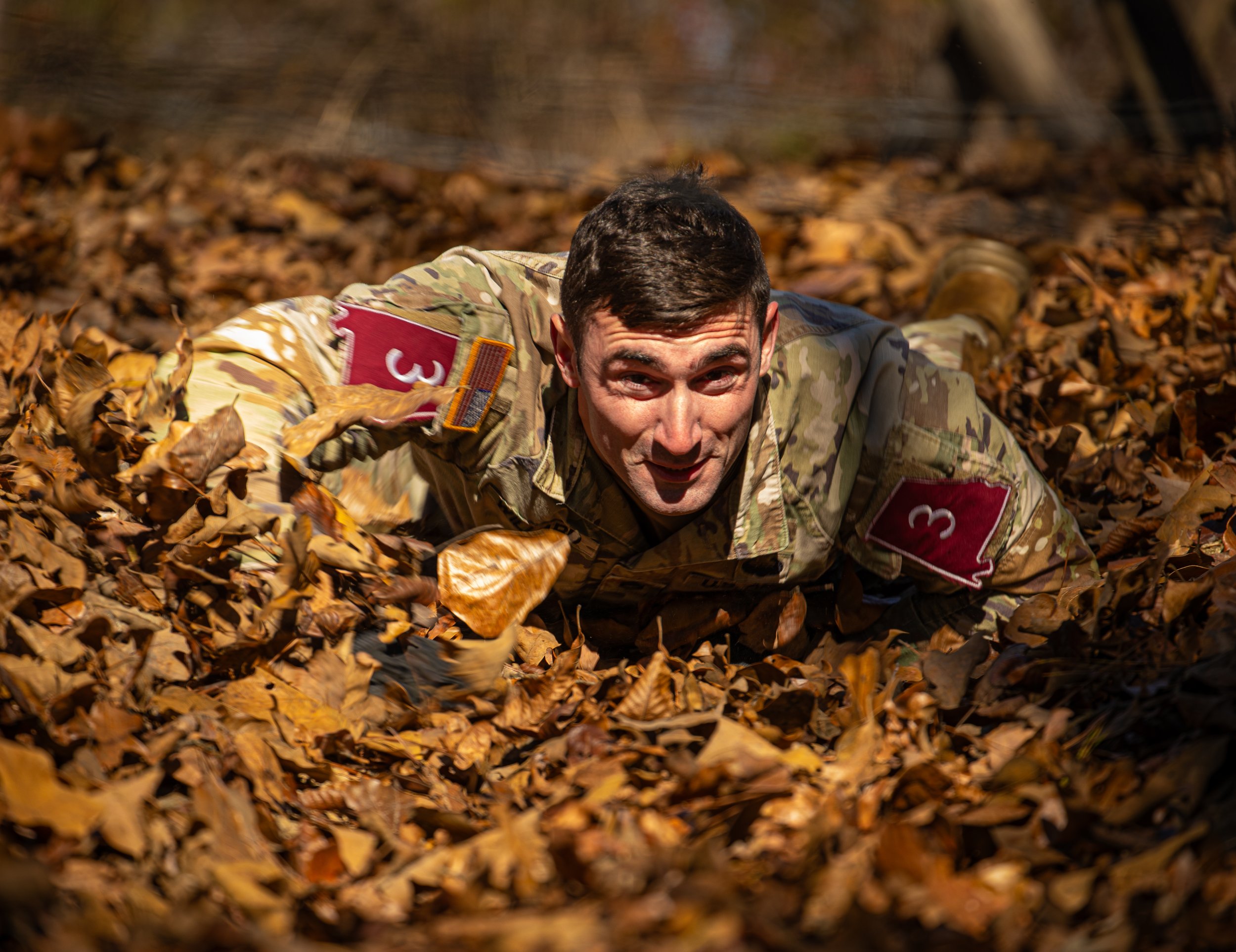 A man in a camouflage military uniform crawling on a bed of fallen autumn leaves.