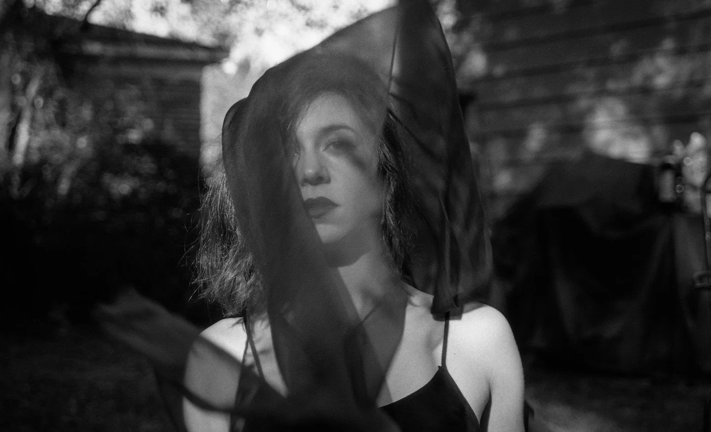 A woman with wavy shoulder-length hair wearing dark makeup and a sheer black veil covering part of her face, standing outdoors near a wooden fence.