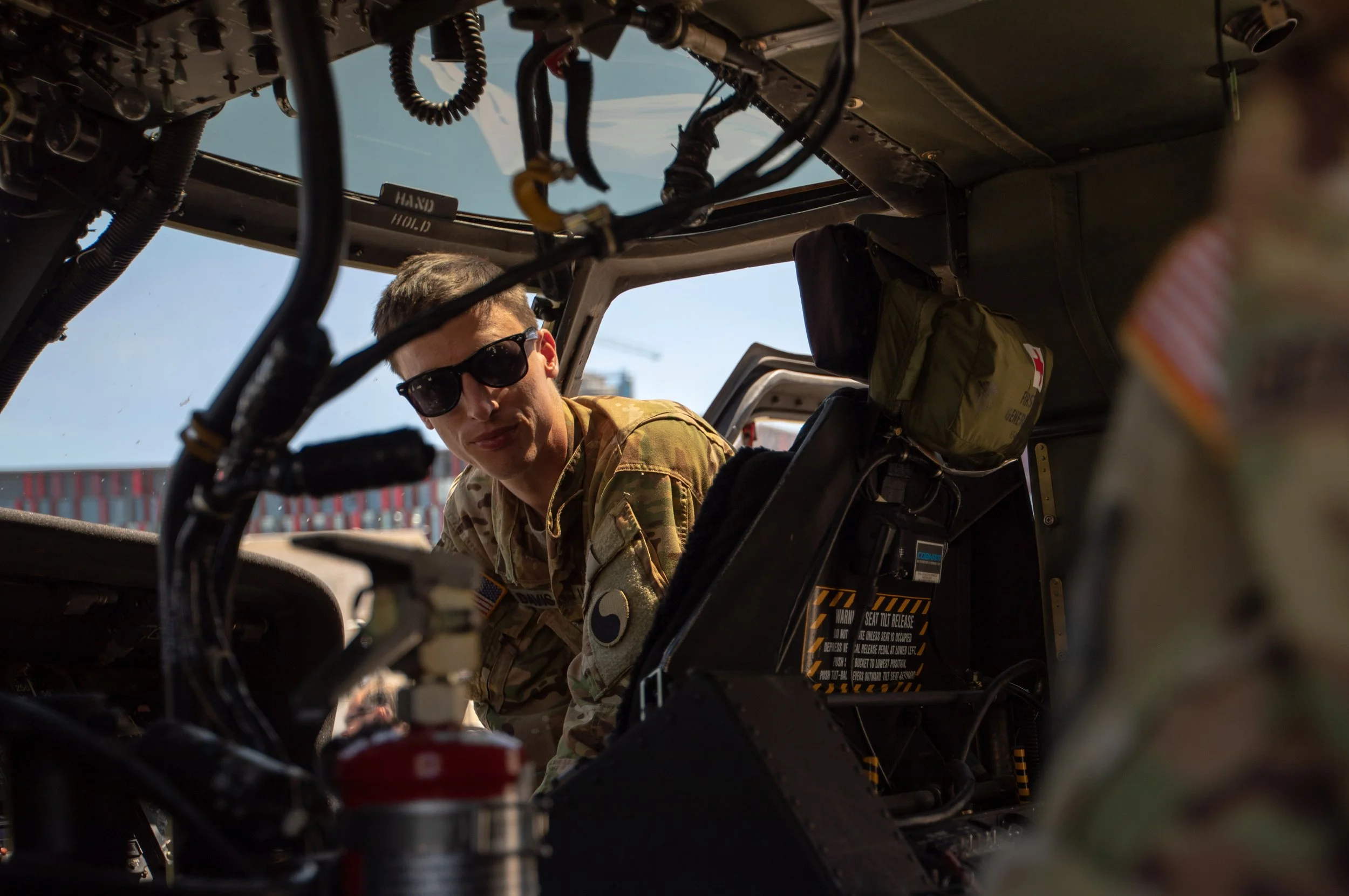 A soldier in camouflage uniform and sunglasses leaning into the cockpit of a helicopter, surrounded by various wires and equipment inside the aircraft.