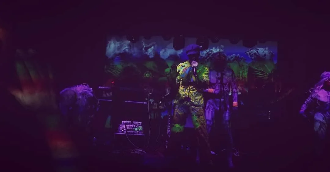 A dark stage with performers in camouflage attire, some wearing hats, and a colorful, abstract background projection.