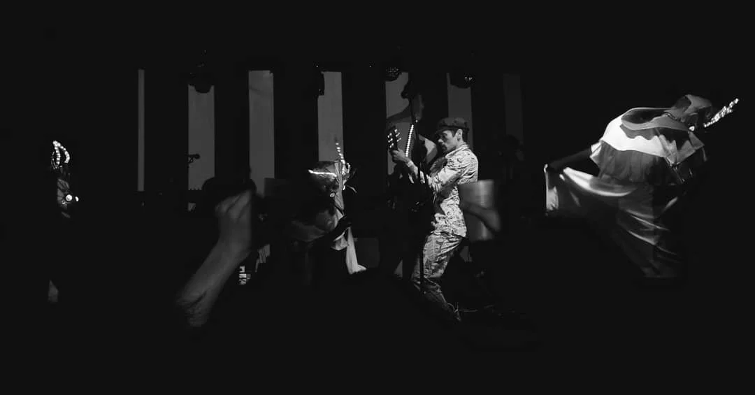 Black-and-white photo of musicians performing on stage, with audience members visible in the foreground.
