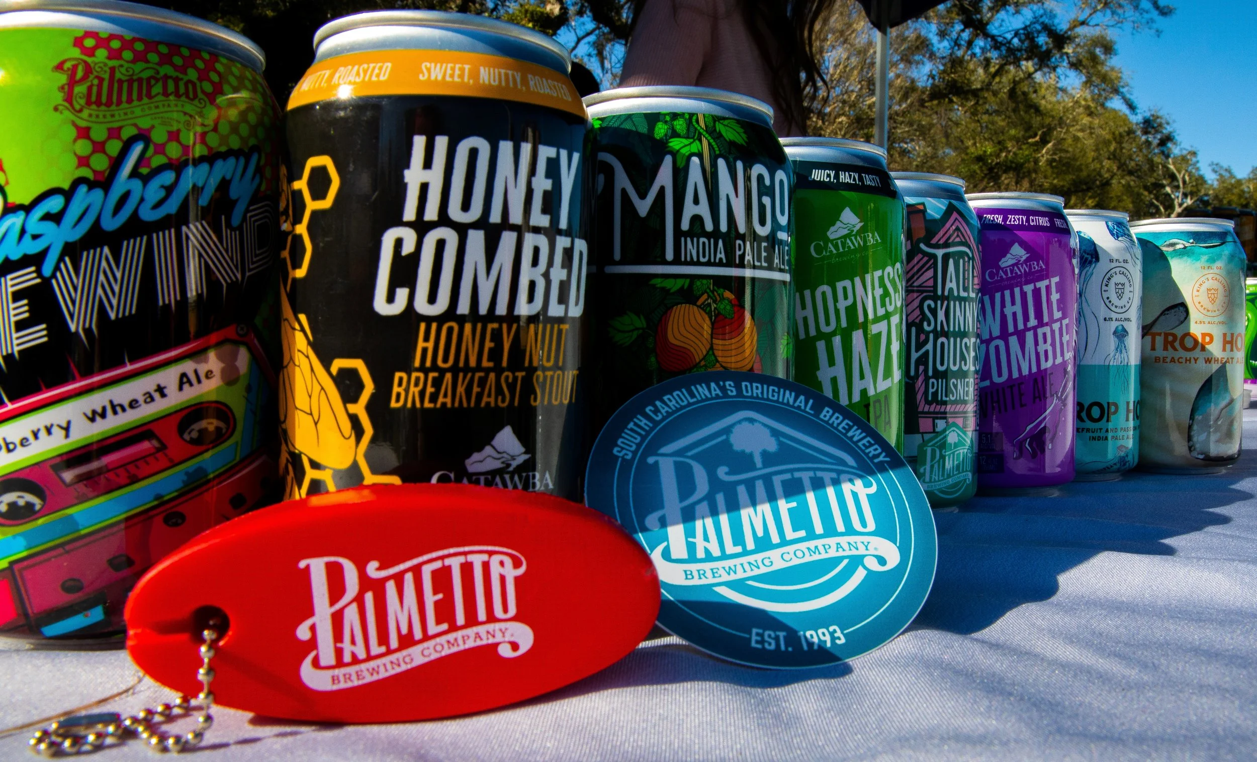 A row of colorful canned beverages on a table, including flavors like cranberry, honey caramel, mango, hopsness haze, white zombie, and Trop Hop Beachy Wheat Ale, with some can labels partially visible and a few round branded tags or coasters in the 