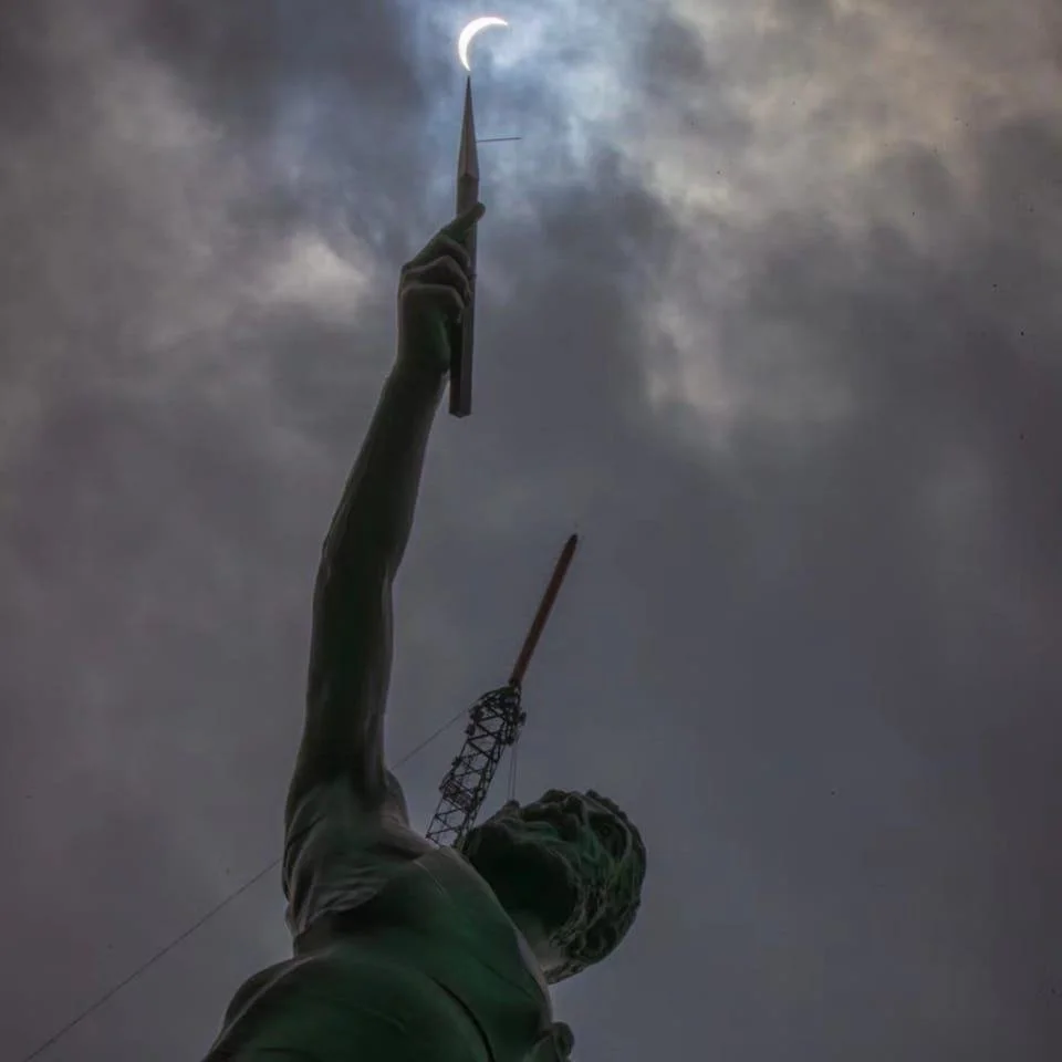 A statue of a woman with an arm raised, pointing a stylus or writing instrument towards the moon in a cloudy sky at night, with a construction crane in the background.