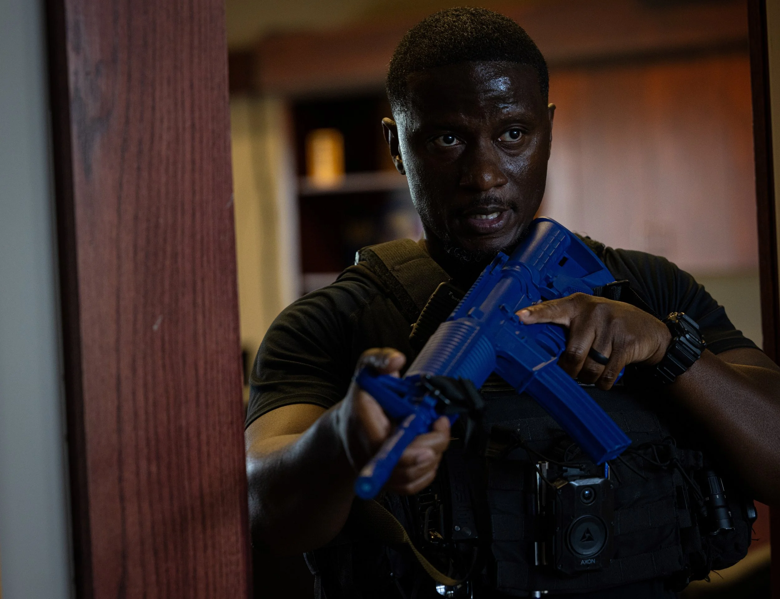 A man with dark skin and short dark hair aiming a blue toy gun around a corner inside a room.