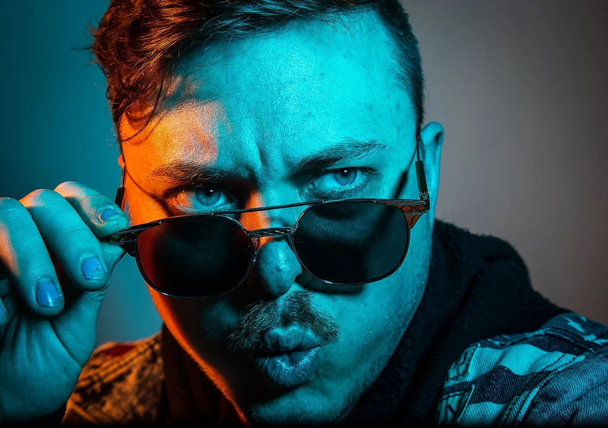A man with blue eyes, partially hidden behind sunglasses, making a surprised or curious expression, with colorful lighting emphasizing his face.